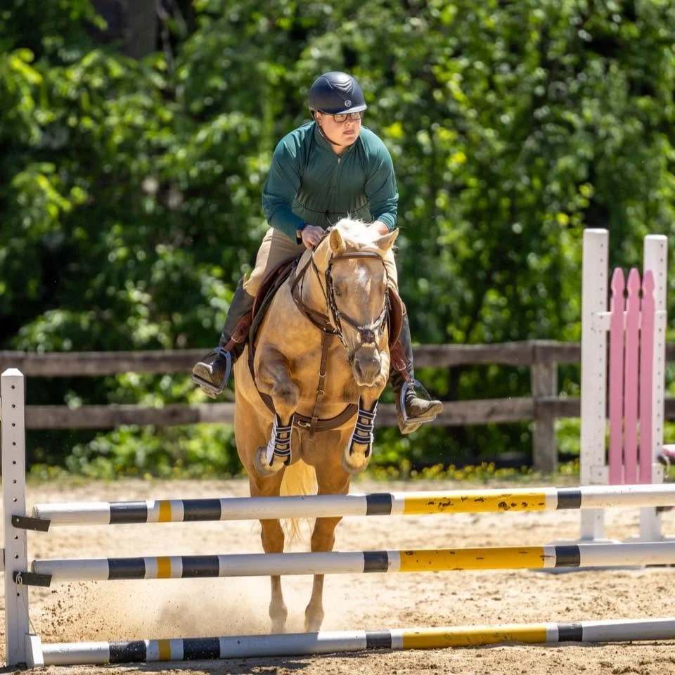 A man riding a palomino horse and jumping over an obstacle.