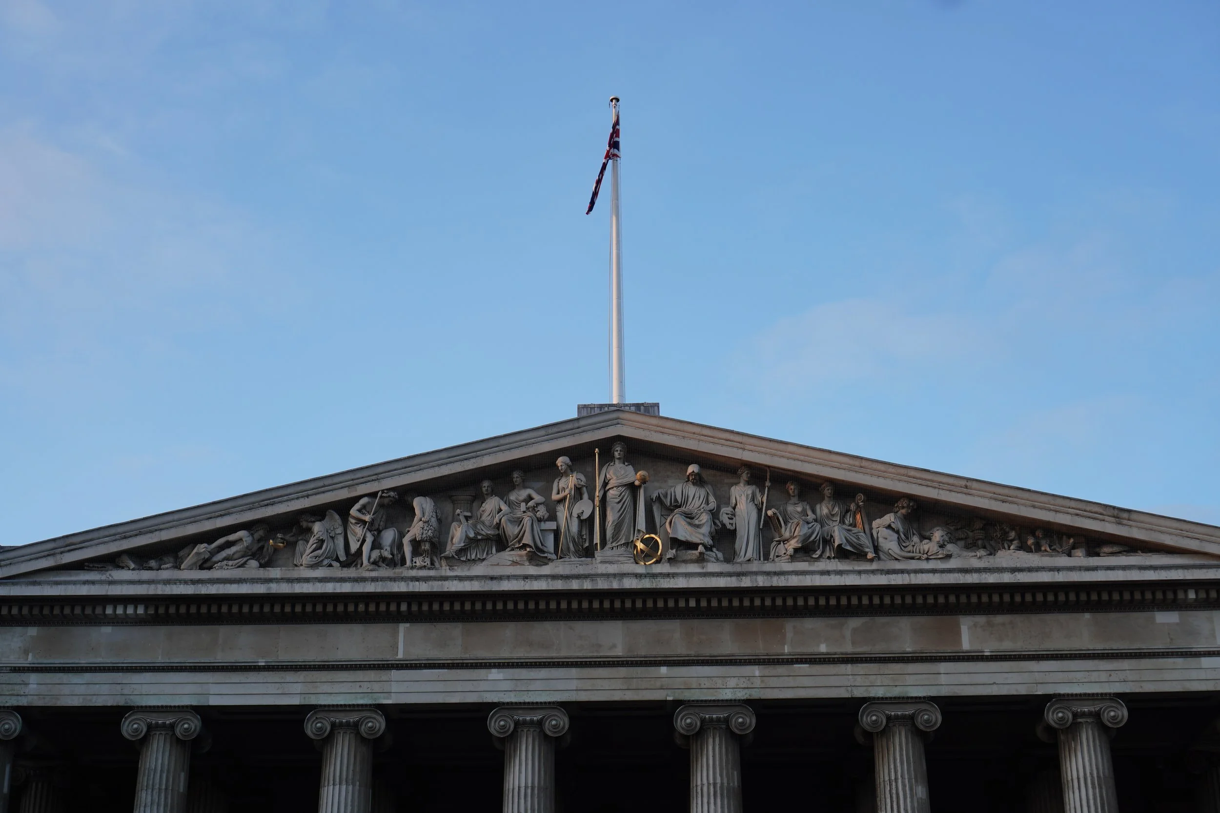 The upper part of the british meuseum with a detailed marble frieze depicting human figures and mythological scenes, and a flagpole with a british flag against a blue sky.