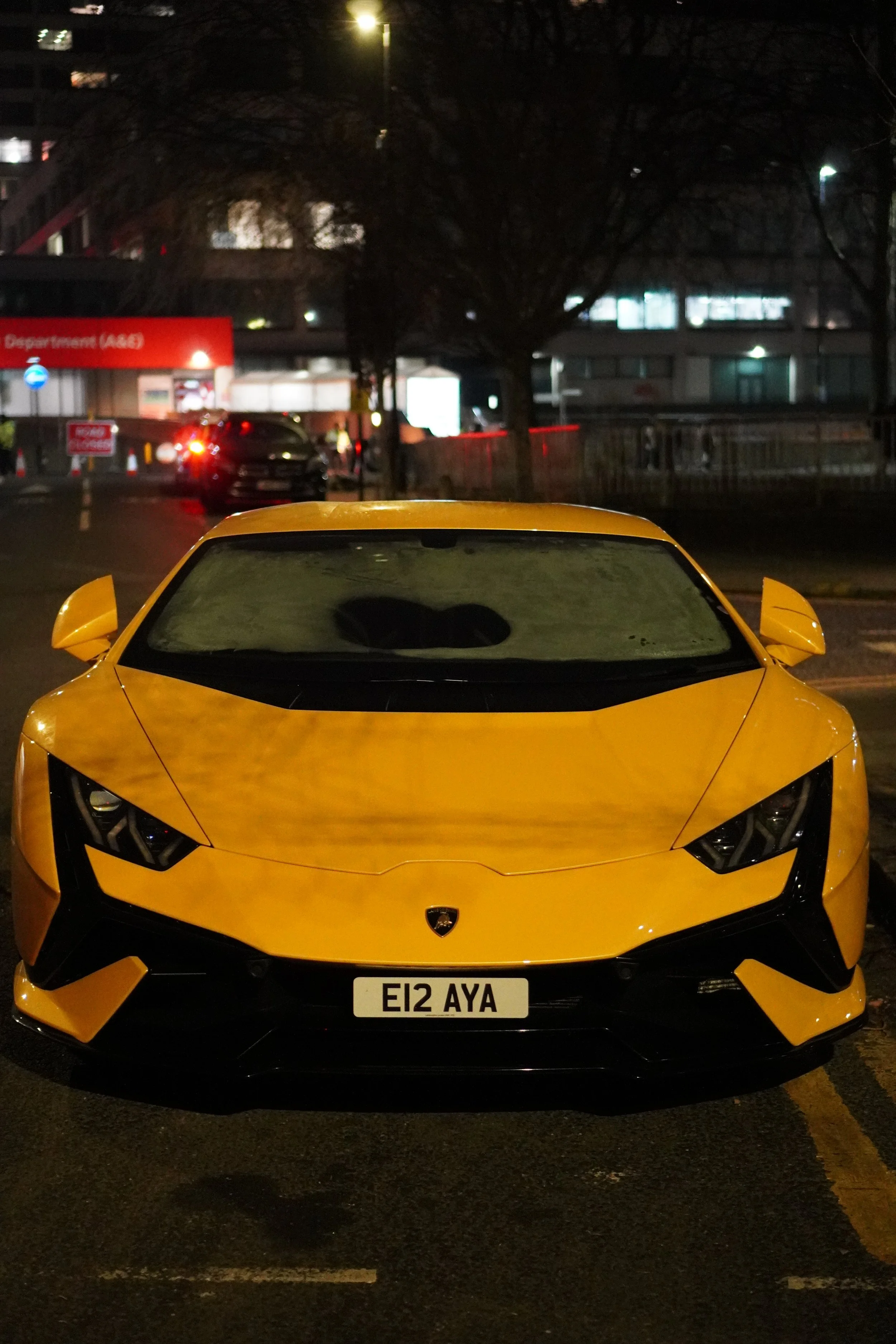 A yellow Lamborghini supercar parked in a dark parking lot at night with a frosted windshield and a custom license plate reading 'E12 AYA'.
