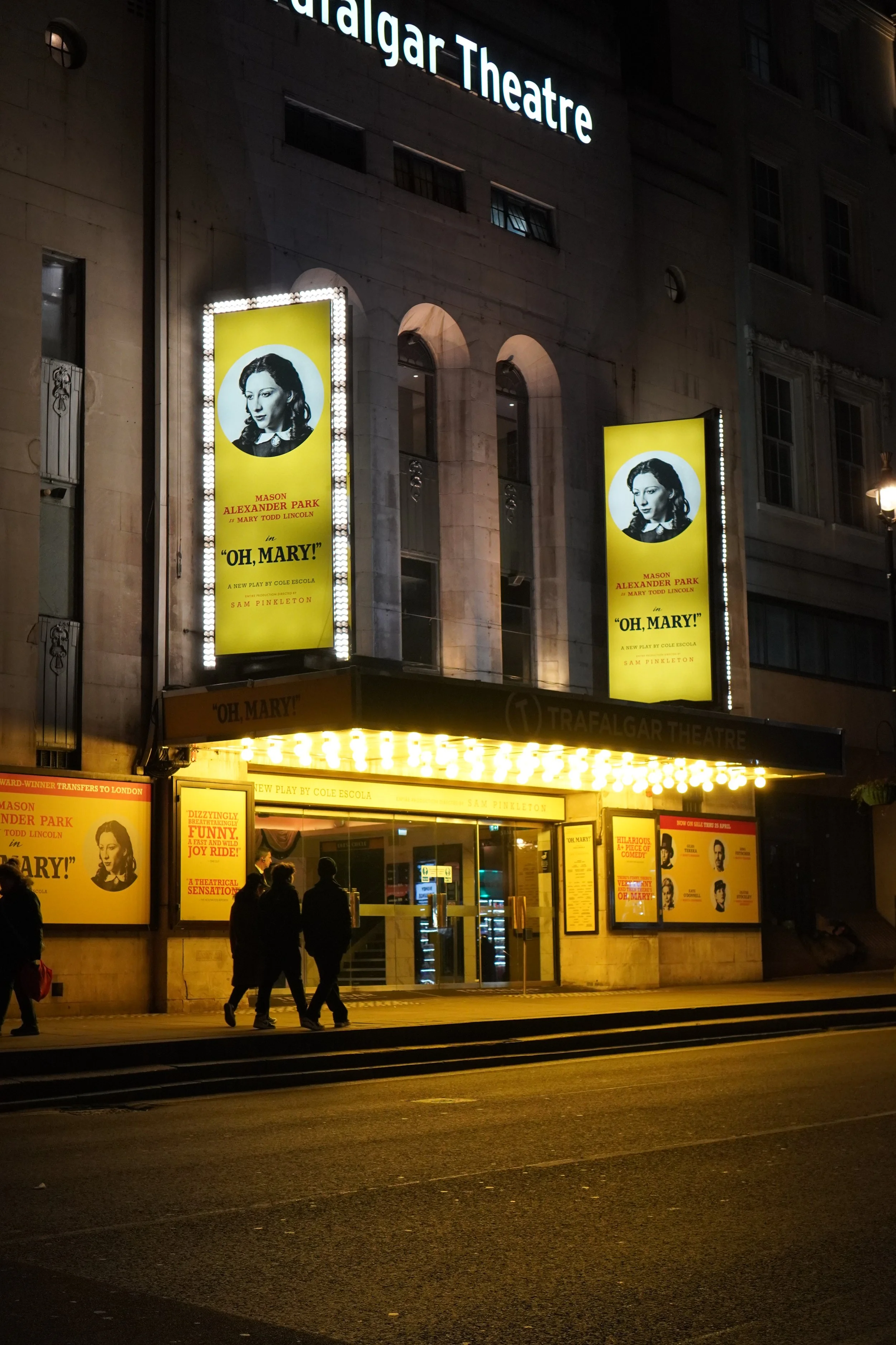 Night view of the Trafalgar Theatre with large illuminated posters advertising the play "Oh, Mary!" featuring Mason Alexander Park at the entrance. Four people walk on the sidewalk in front of the theatre.