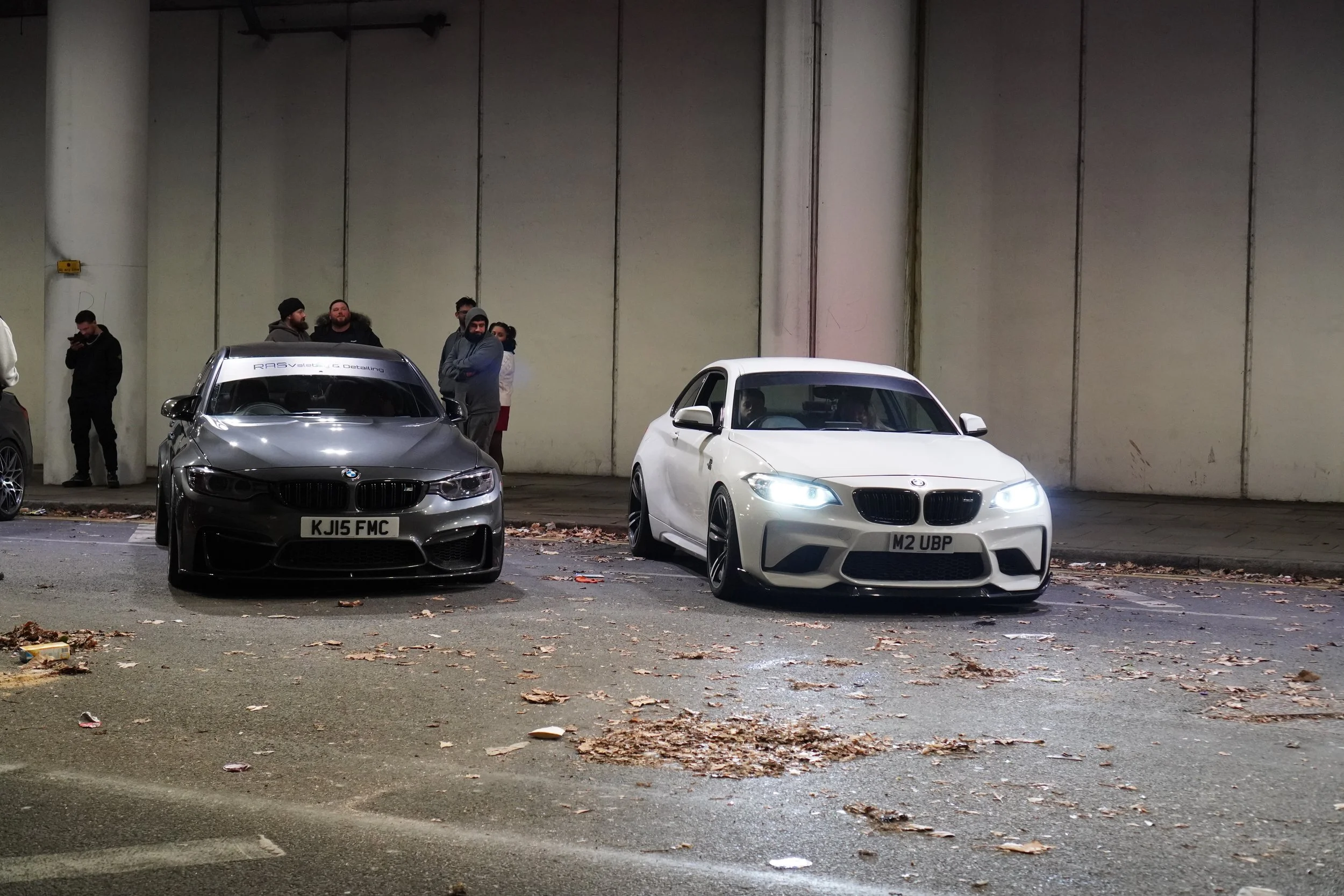 Two BMW cars parked on a street at night, with several people standing in the background.