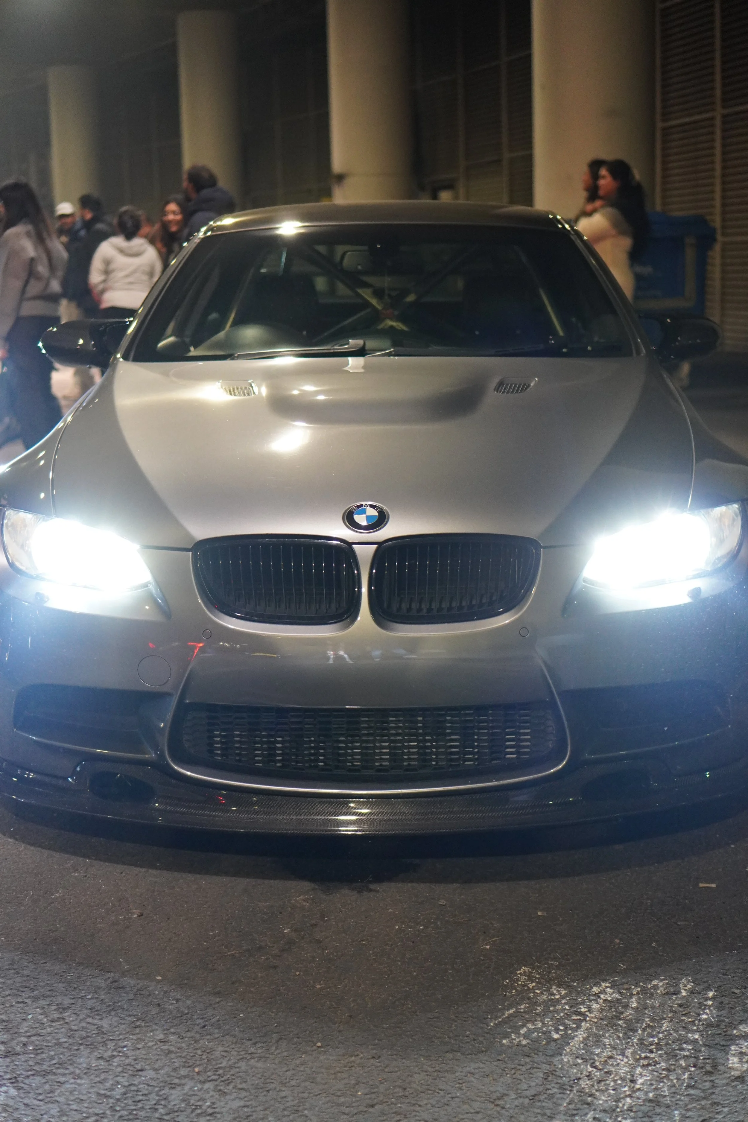 Front view of a silver BMW car with bright headlights turned on at night, surrounded by people.