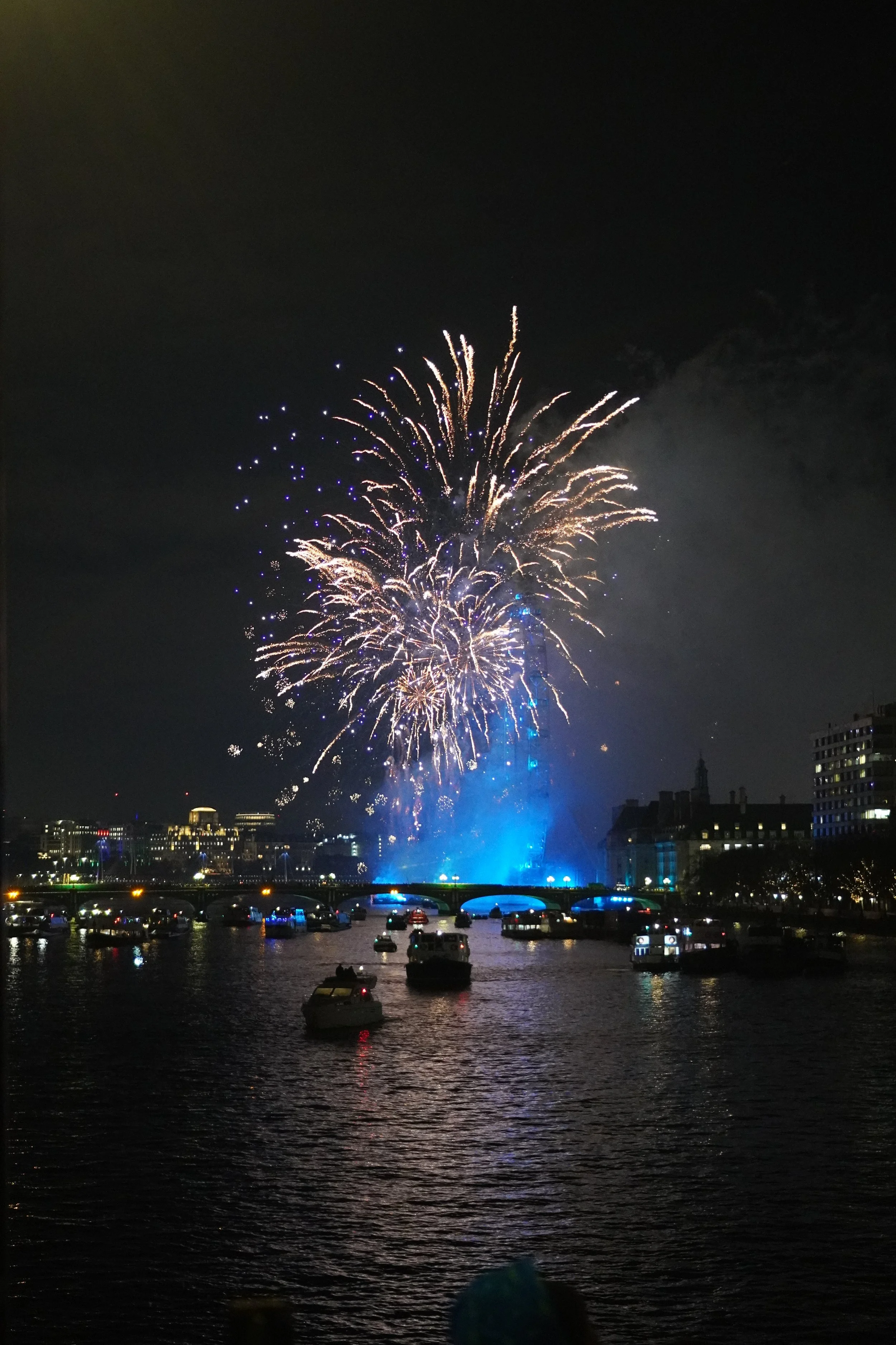 Fireworks display on new years eve on a bridge near London Eye with boats in the water and city buildings in the background.