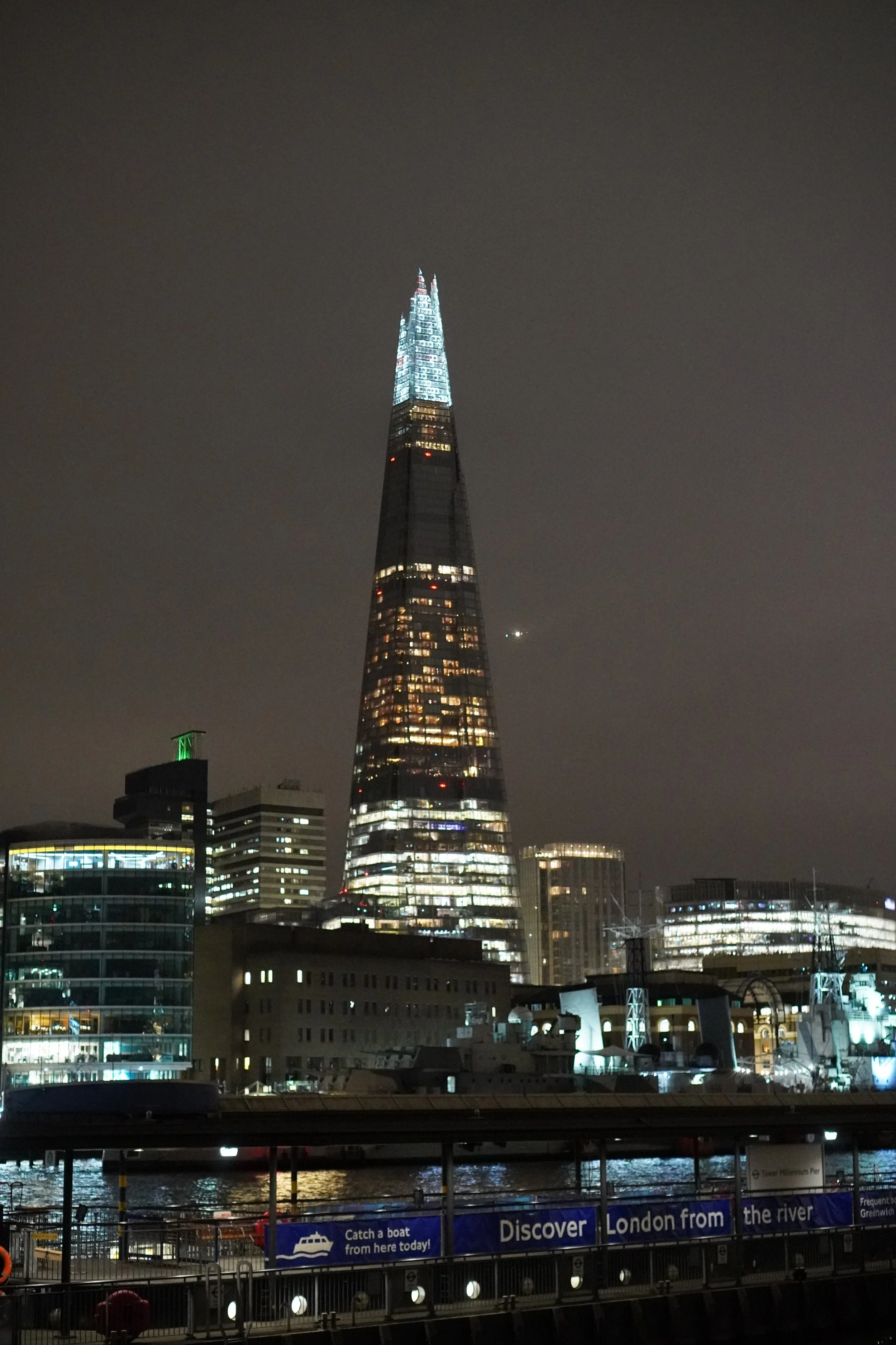 Nighttime view of The Shard skyscraper illuminated with blue lights, surrounded by other buildings and a river in London.