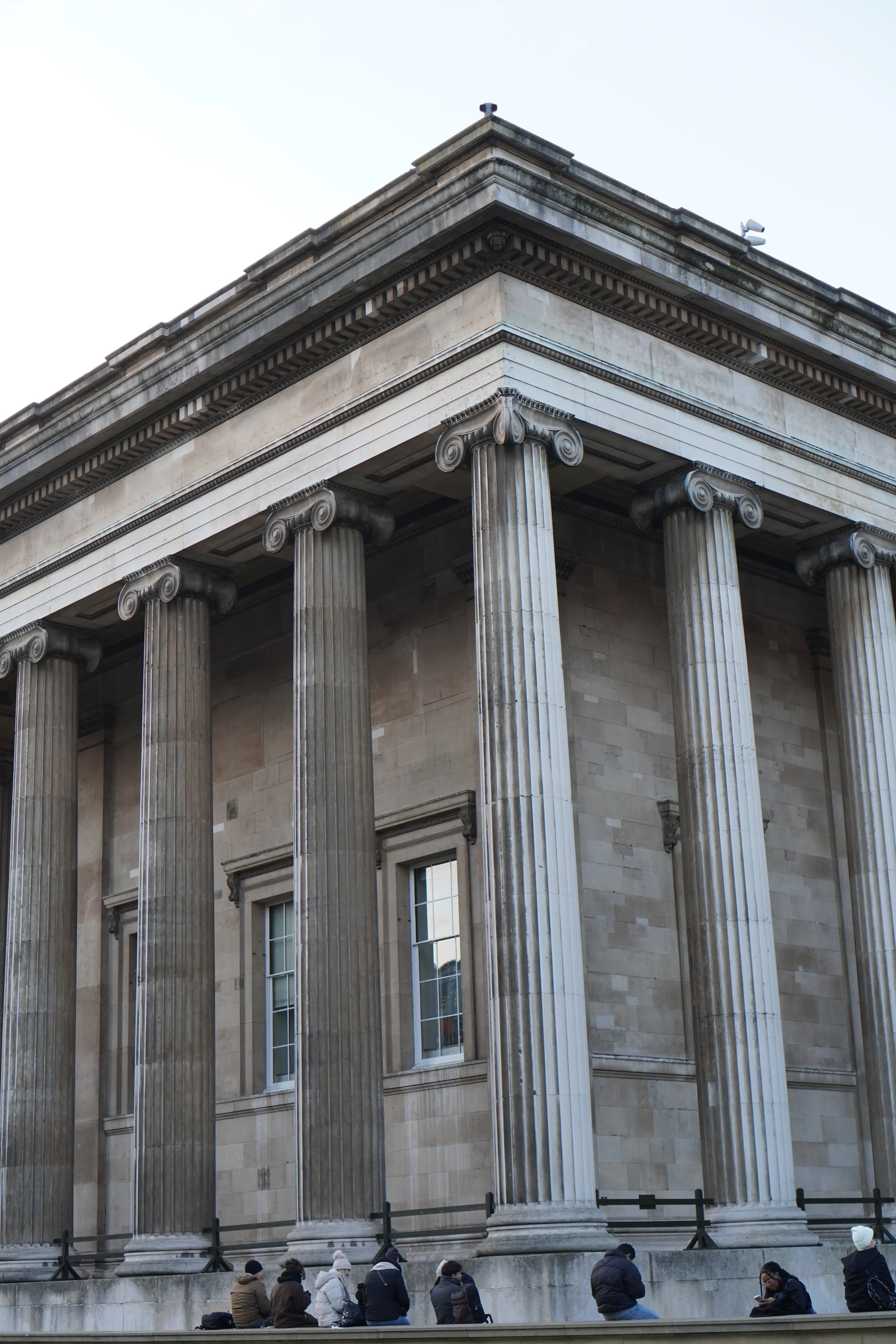 People sitting on a stone platform outside of the British meuseum with tall, fluted columns and ornate capitals.