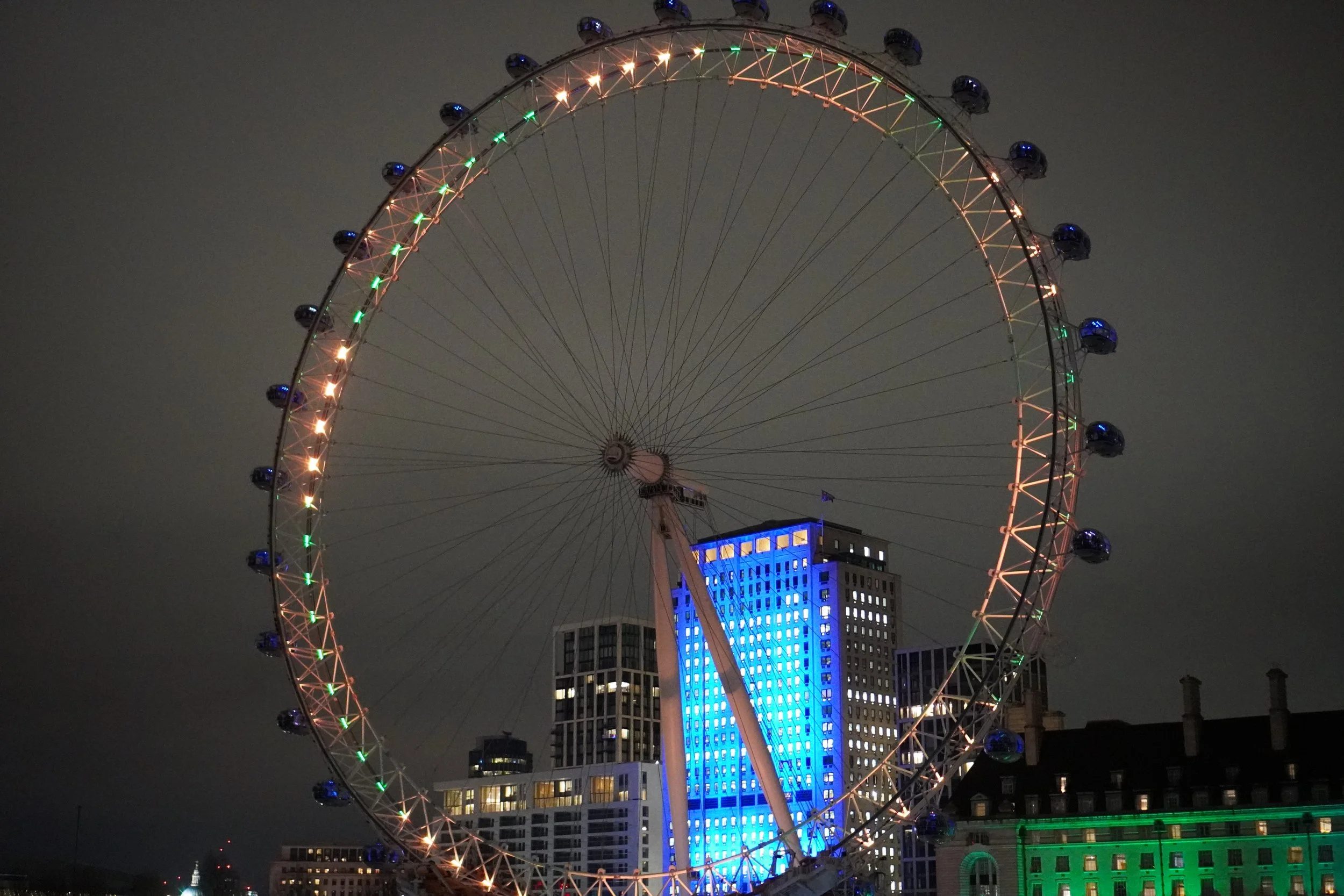 Night view of a lit-up London with colored lights against a city skyline, some buildings illuminated with blue and green lights.