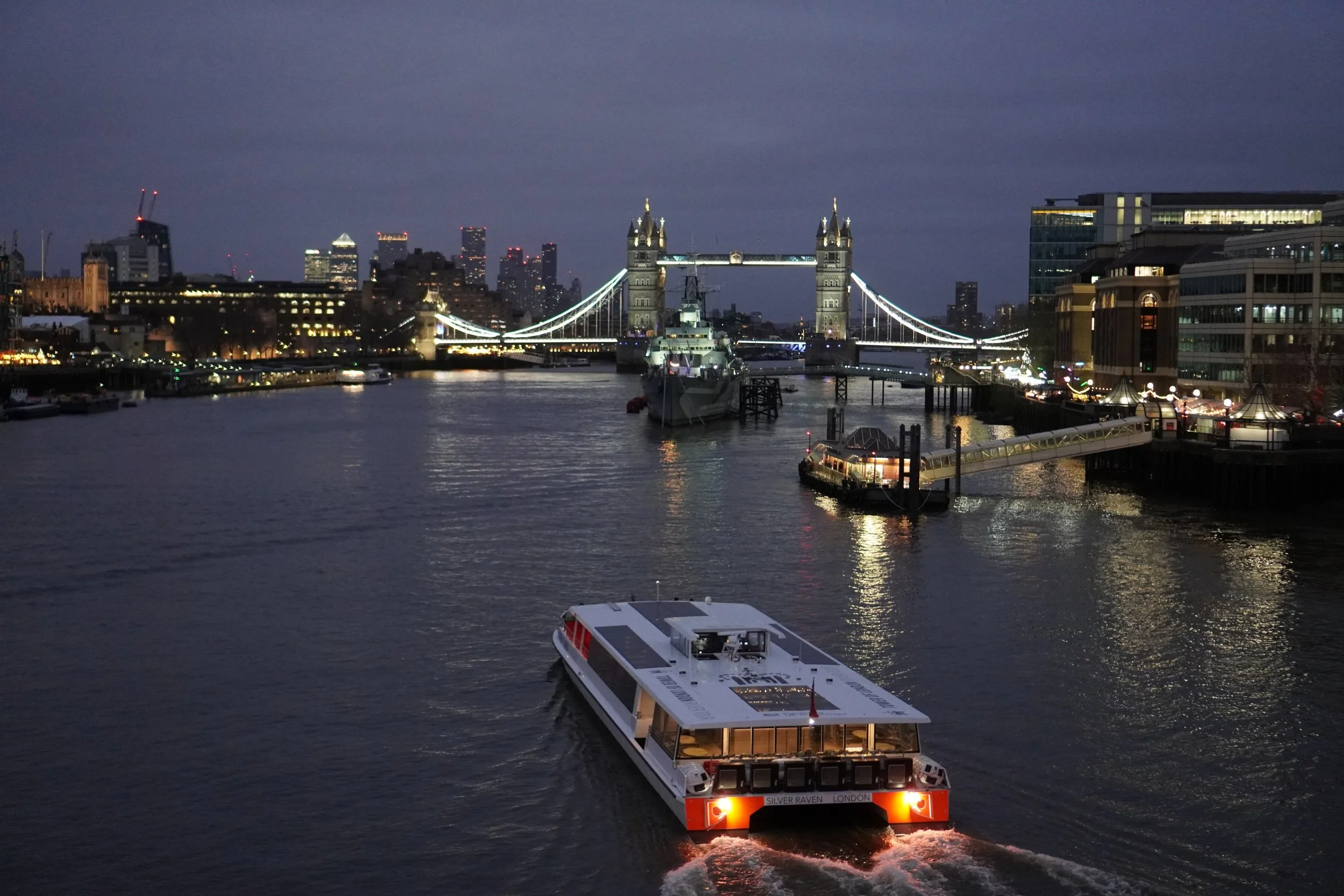 Nighttime view of the London skyline featuring Tower Bridge illuminated over the River Thames, with a boat in the foreground and office buildings in the background.