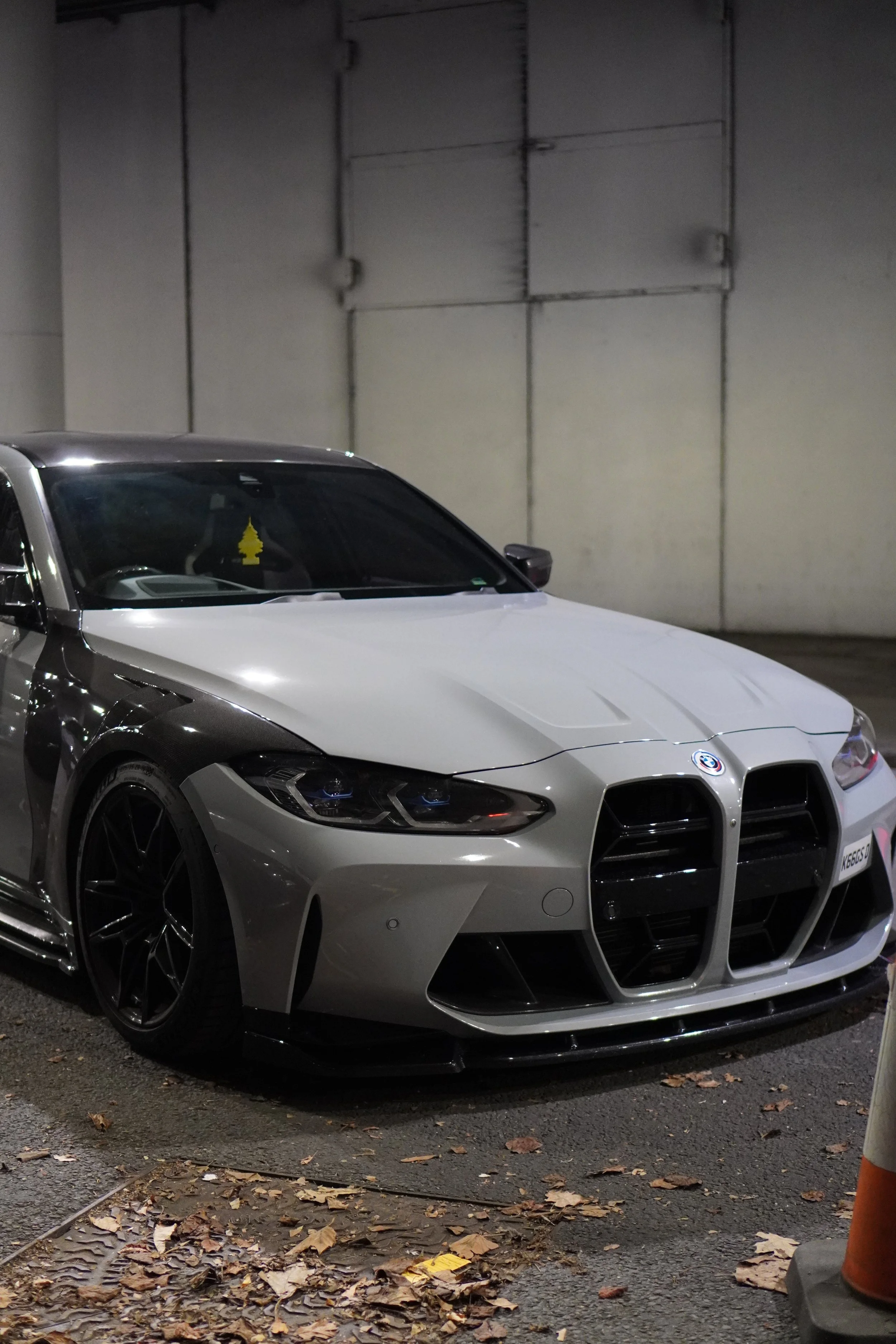 A white BMW car parked in an underground parking lot with a yellow tree-shaped air freshener hanging from the rearview mirror. The ground is scattered with dry leaves, and there is a white wall with metal panels in the background.