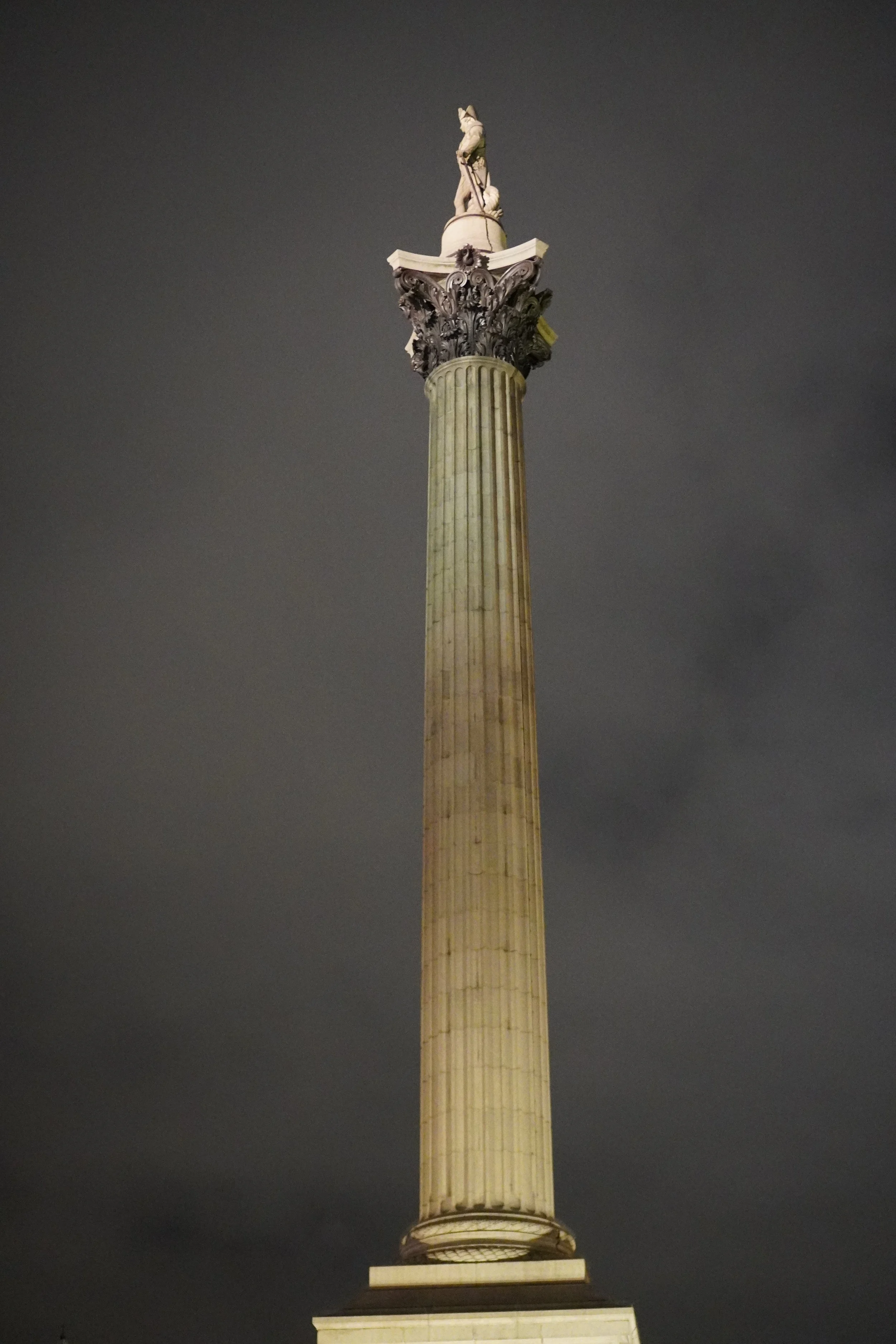 Night view of a tall, illuminated classical-style stone column with a statue on top, against a cloudy dark sky.