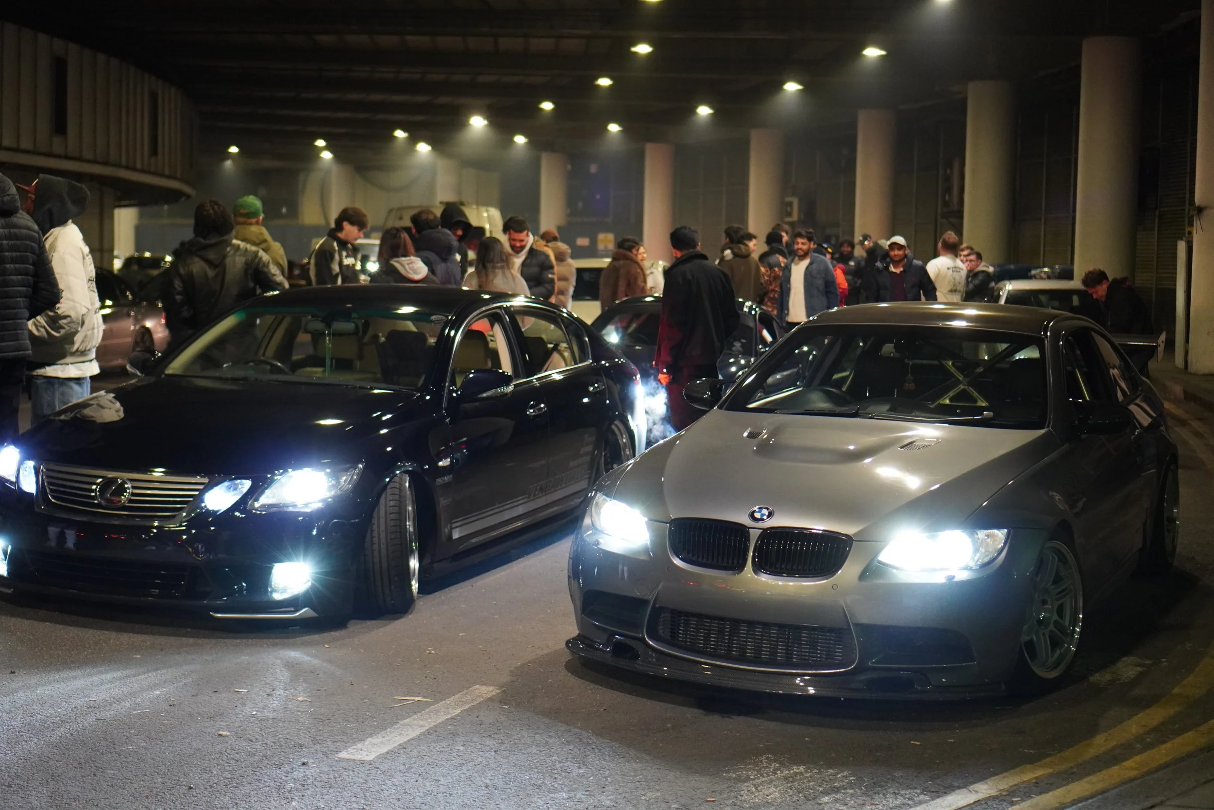 Nighttime indoor car meet with people gathered around two lowered, customized sports cars, a black Lexus and a gray BMW, inside a parking garage.