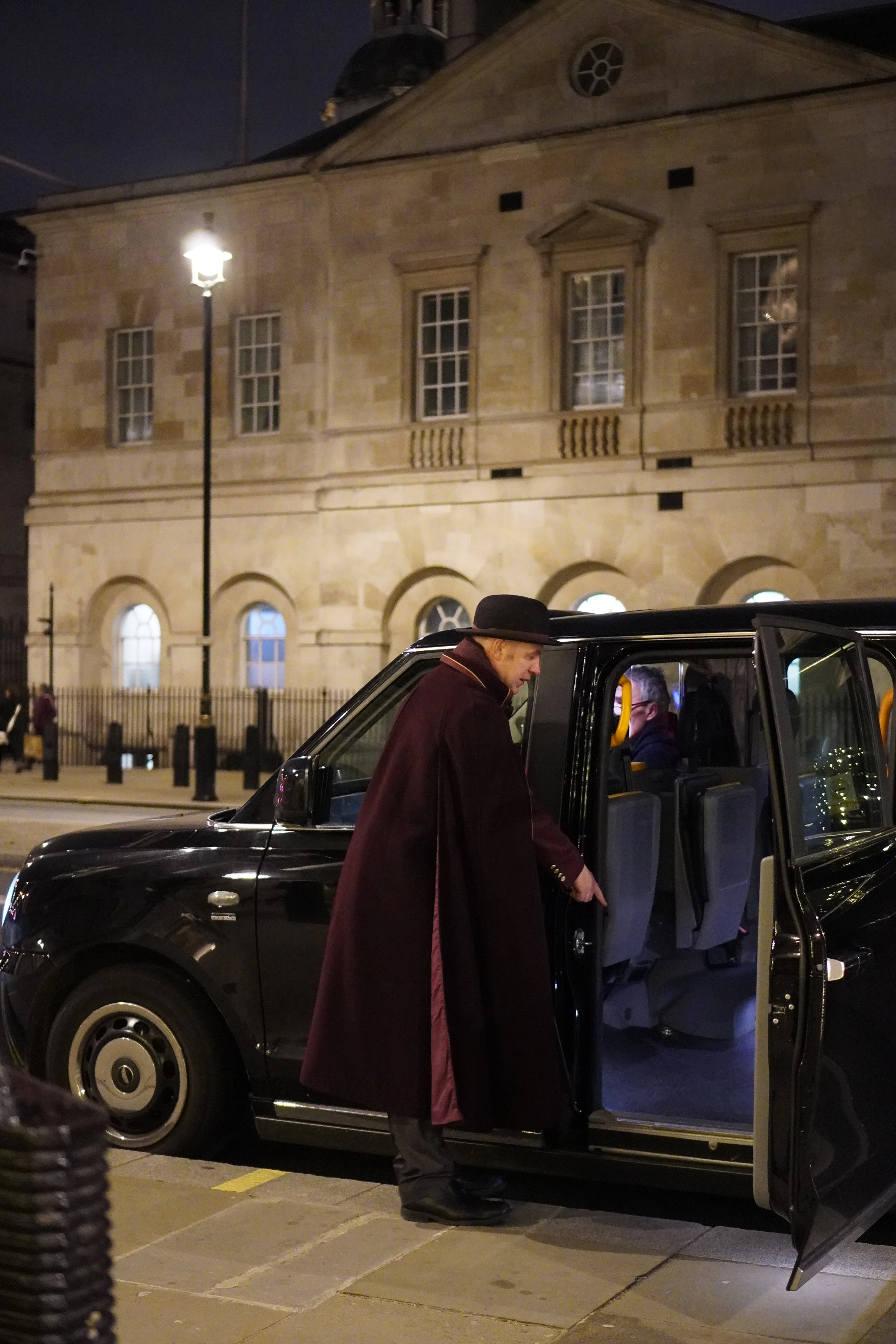 A man in a dark coat and hat boarding a black taxi cab at night, with a historic stone building in the background.