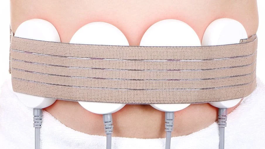 Person wearing a beige abdominal belt with six attached electrodes and wires for medical monitoring.