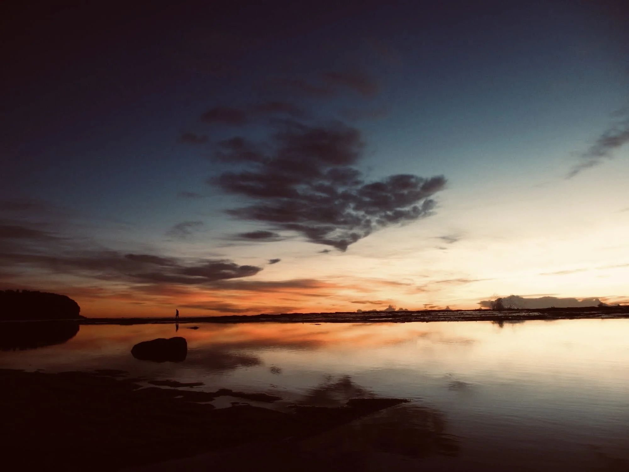 Sunset over a calm ocean with a silhouette of a person walking on the beach, clouds in the sky, and reflections on the water.