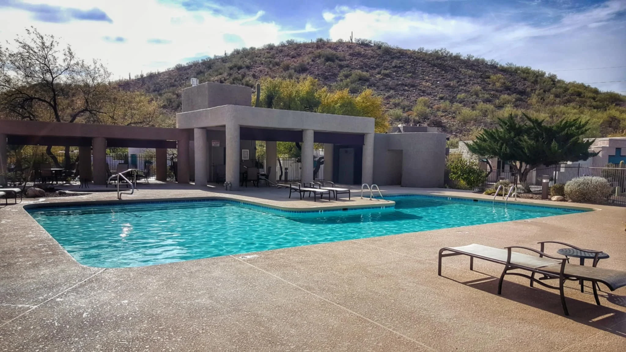 Outdoor swimming pool with lounge chairs and a shaded seating area, surrounded by desert vegetation and a mountain in the background.