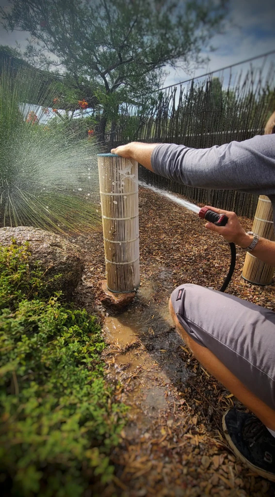 Person watering a tall bamboo fountain with a garden hose in a backyard garden.