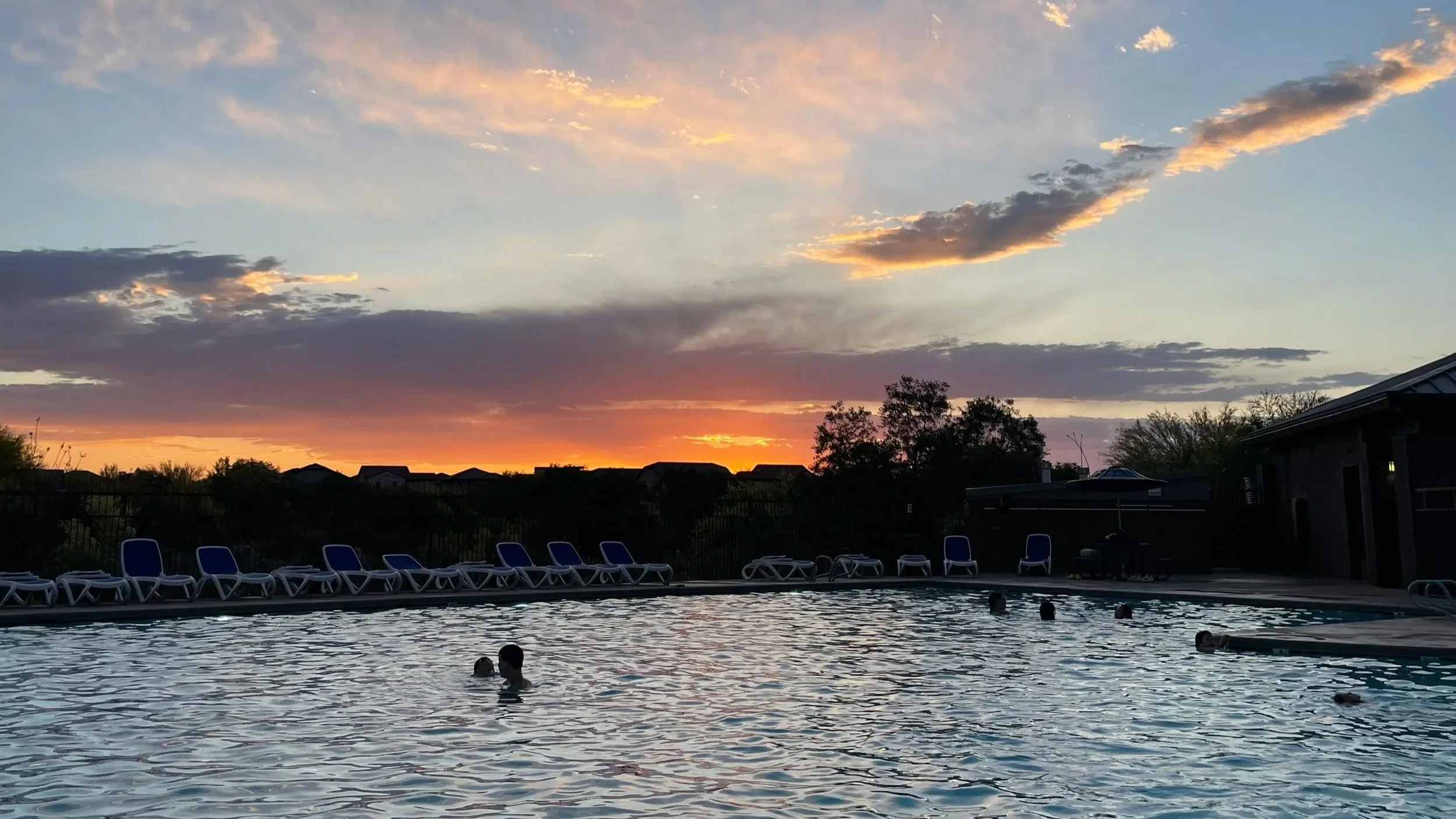 People swimming in an outdoor pool at sunset, with lounge chairs along the poolside and a colorful sky with clouds.