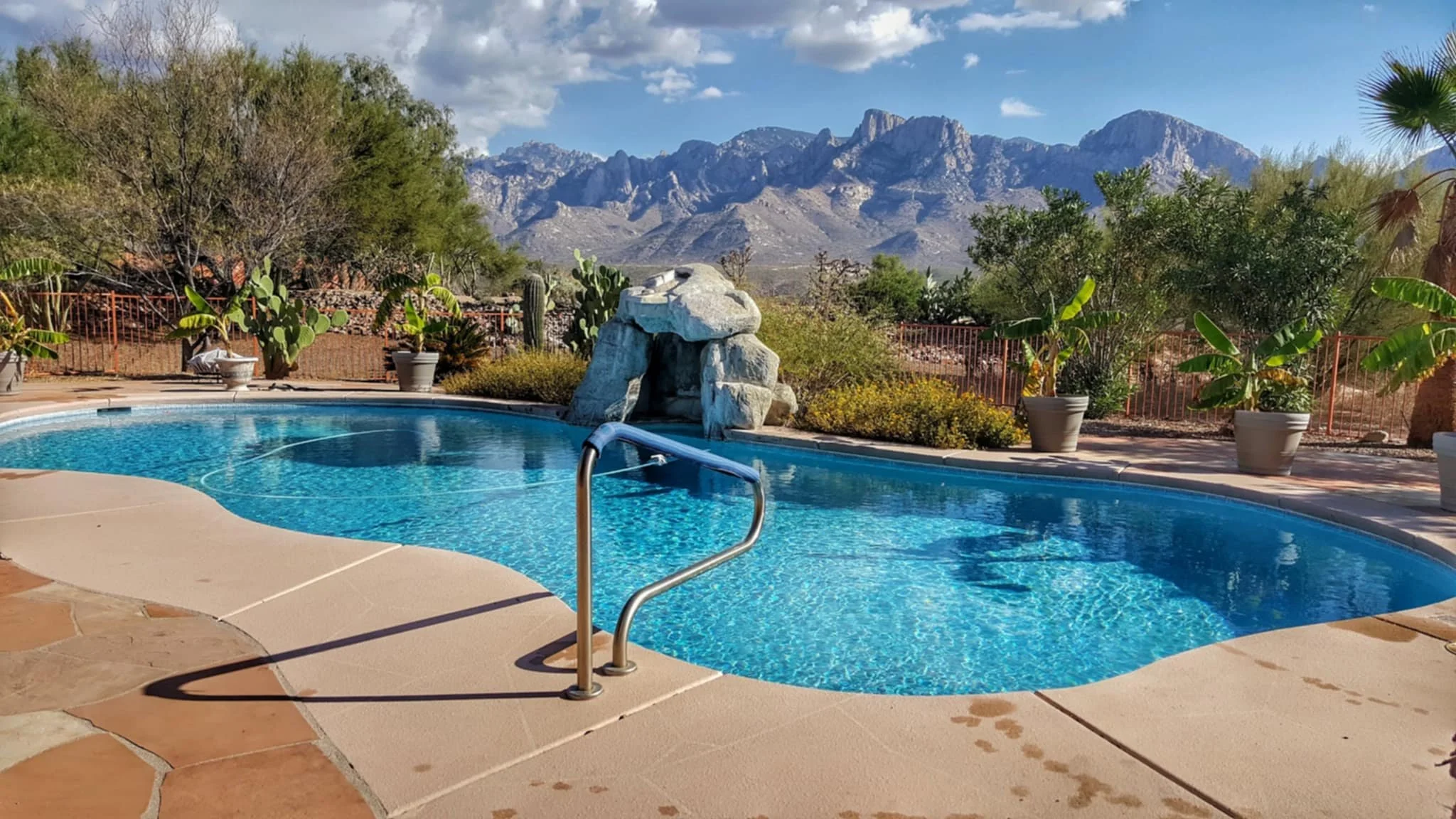 A backyard swimming pool with a rocky waterfall feature, surrounded by potted plants, palm trees, and desert shrubs. Mountain range visible in the distance under a partly cloudy sky.