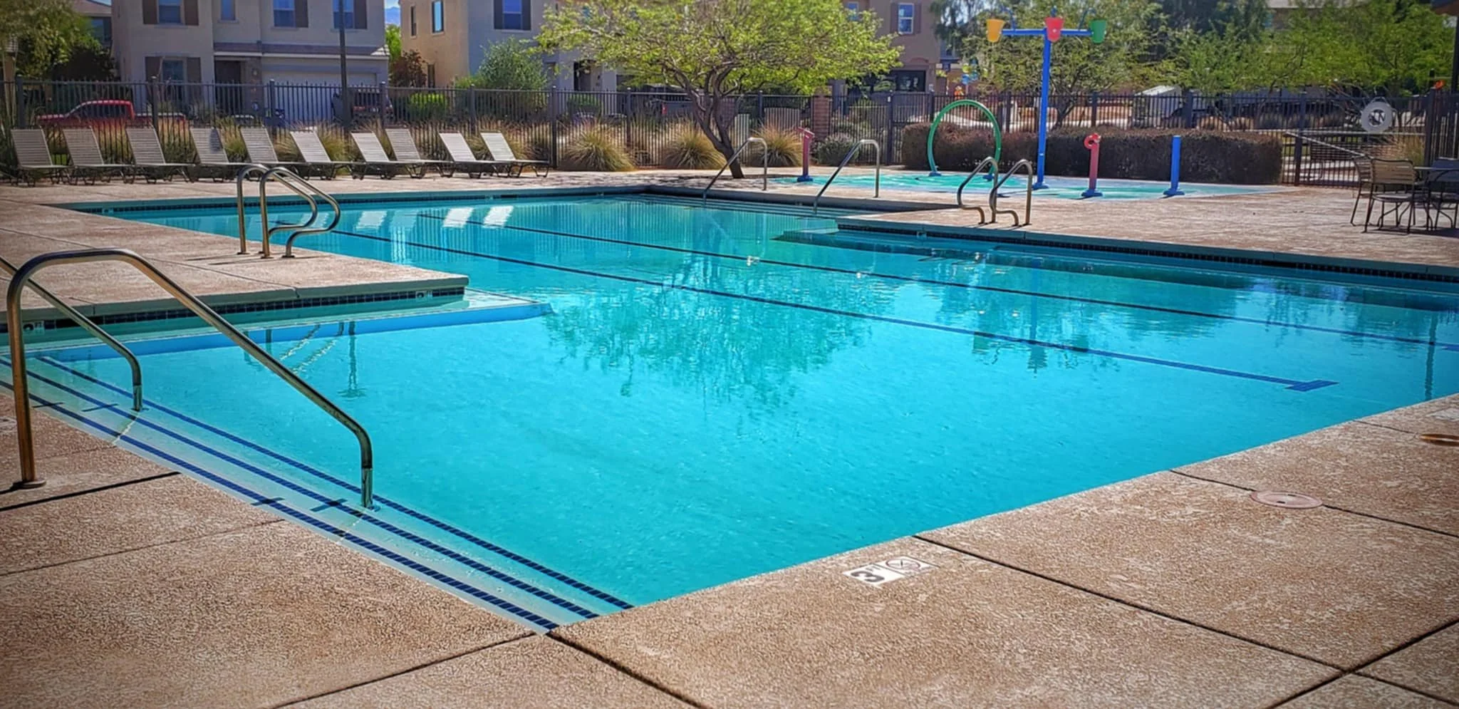 Empty outdoor swimming pool with metal handrails and steps, surrounded by lounge chairs, in a residential complex with trees and apartment buildings in the background.