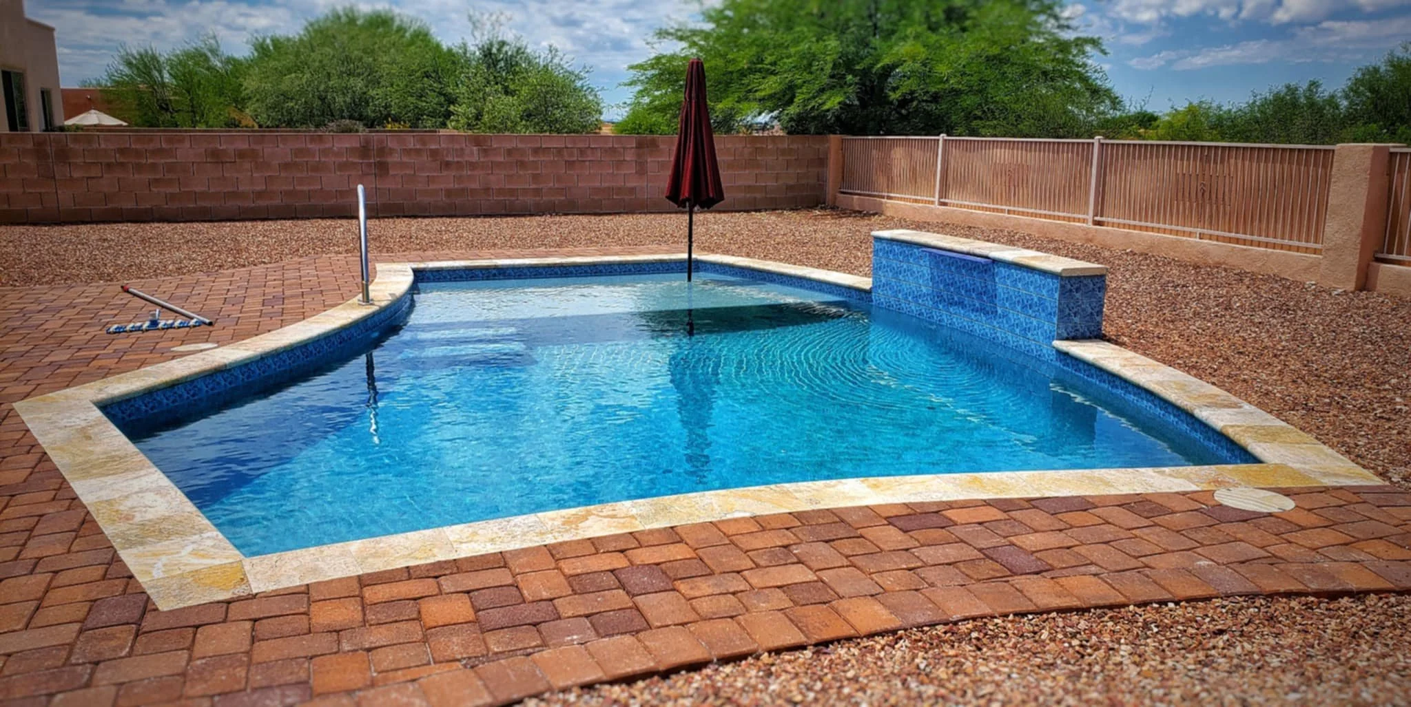 Backyard swimming pool with blue water, a set of steps, and a small waterfall feature, surrounded by brick pavers and a gravel yard, enclosed by a wooden fence and brick wall, with trees and a partly cloudy sky in the background.