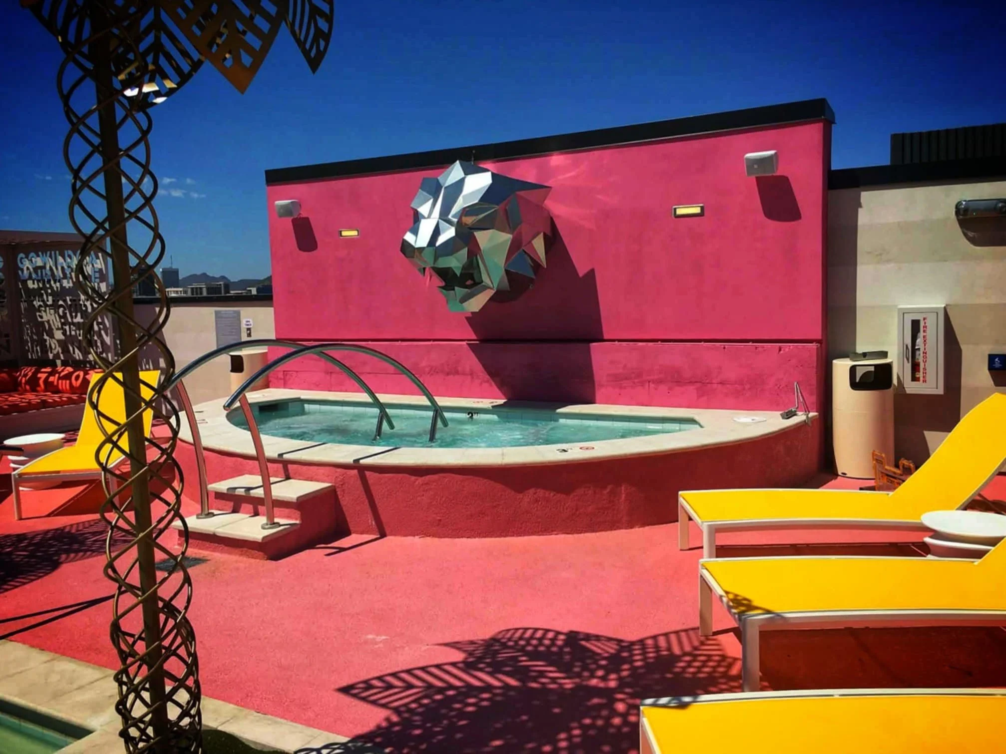 A rooftop pool area with yellow lounge chairs, a pink wall with a geometric lion sculpture, and a small pool with steps and handrails, under a clear blue sky.