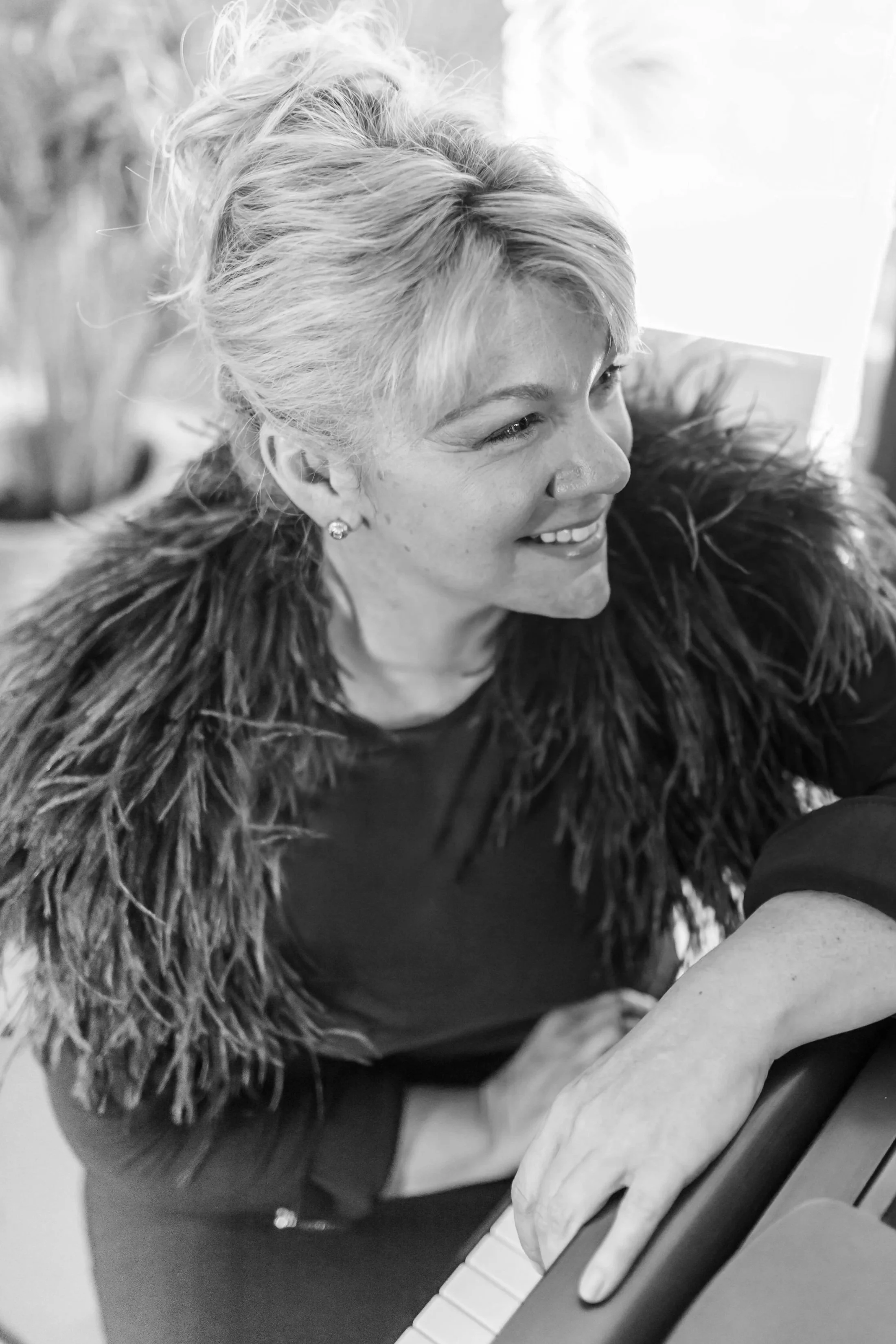 A woman with short, wavy, blonde hair wearing earrings and a fur coat, playing the piano.