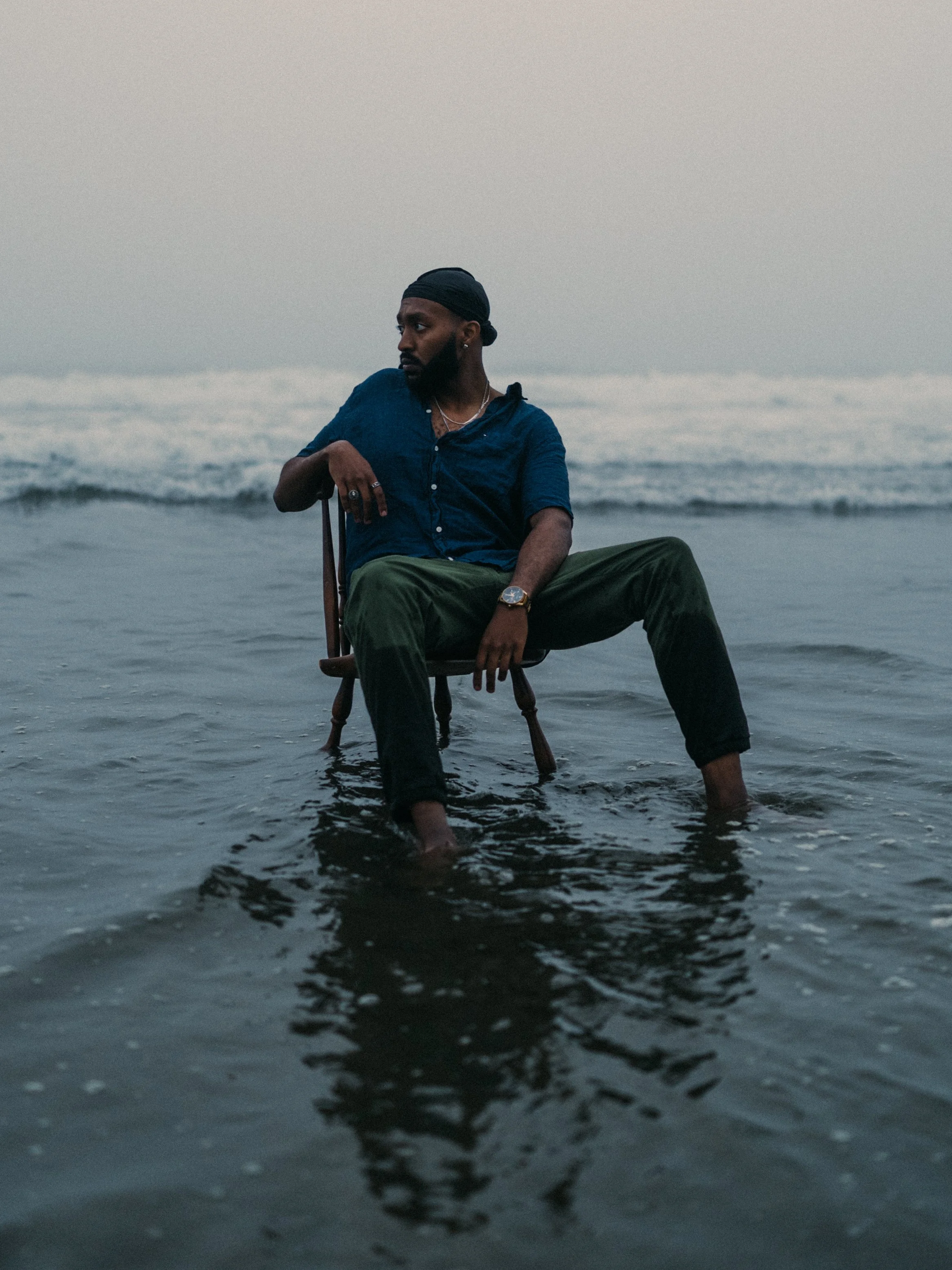 A man sitting on a wooden chair with his feet submerged in shallow water at the beach during dusk.