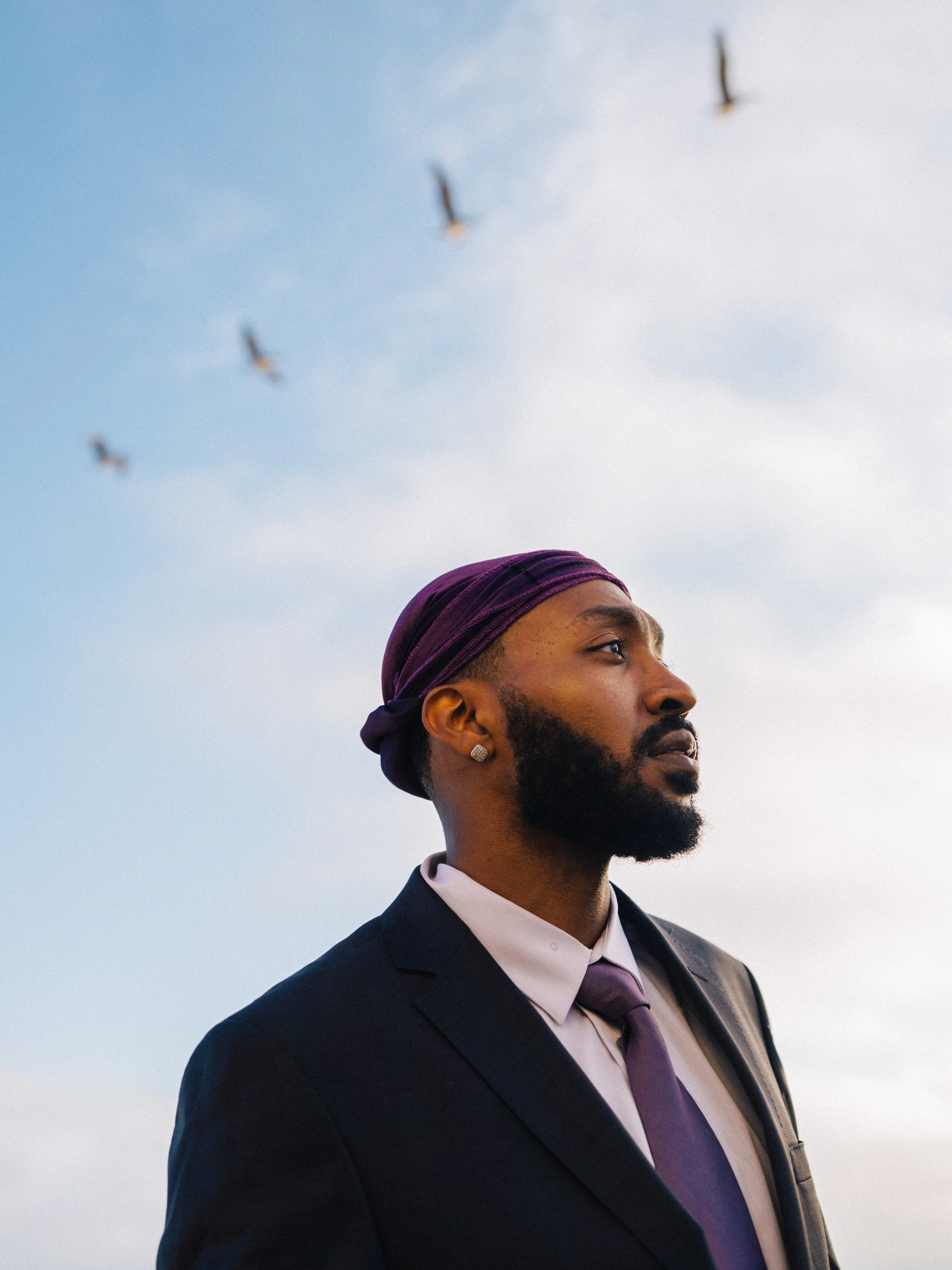 A man with a beard and earring wearing a suit and purple headscarf looking up towards the sky with birds flying in the background.