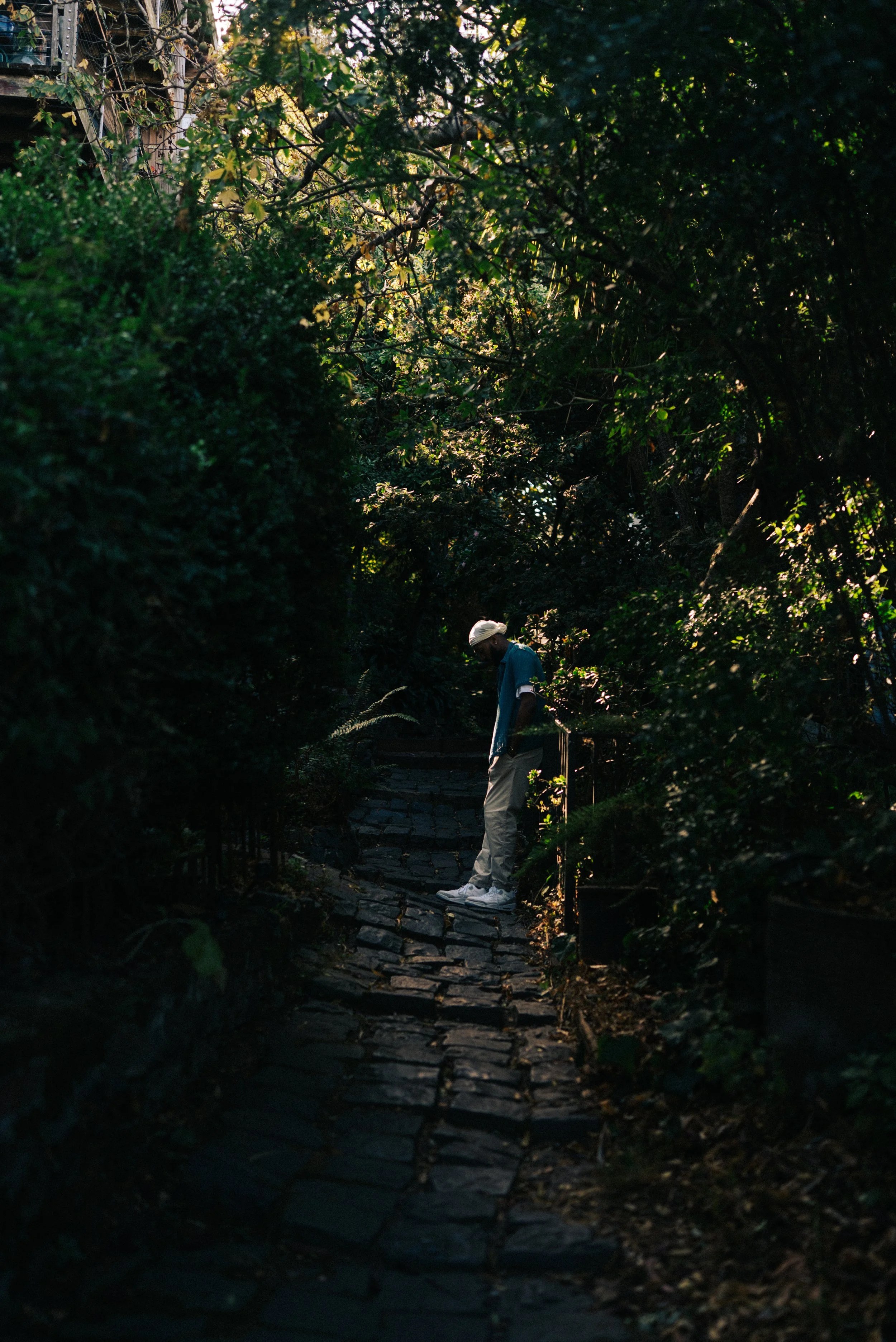 A person with a white cap, blue shirt, and light-colored pants standing on a narrow, cobblestone path surrounded by dense green foliage and trees.