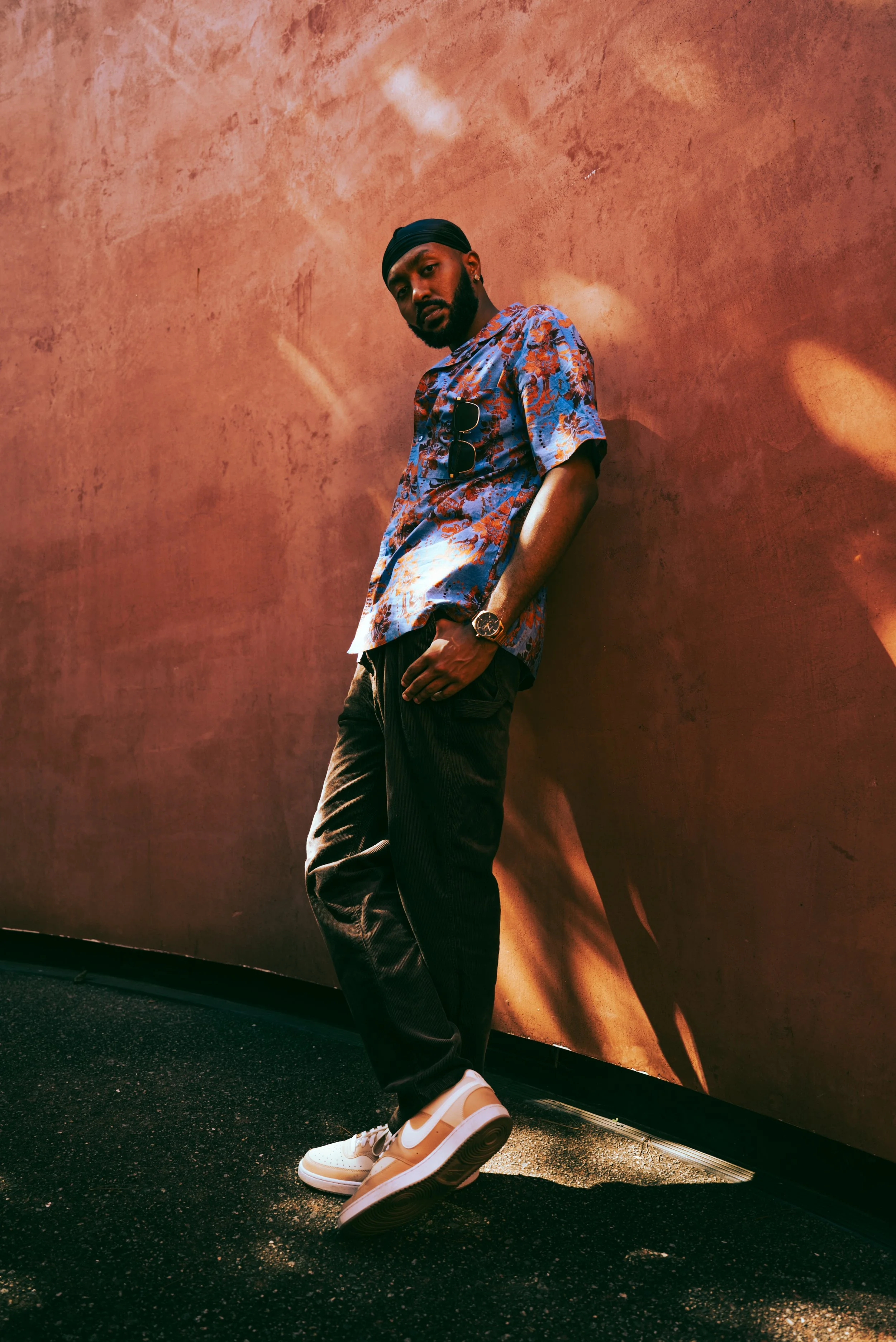A man wearing a black headwrap, blue patterned shirt with sunglasses hanging from the front, black pants, white Nike sneakers, and a watch, leaning against a reddish-brown wall with shadows and sunlight reflections.