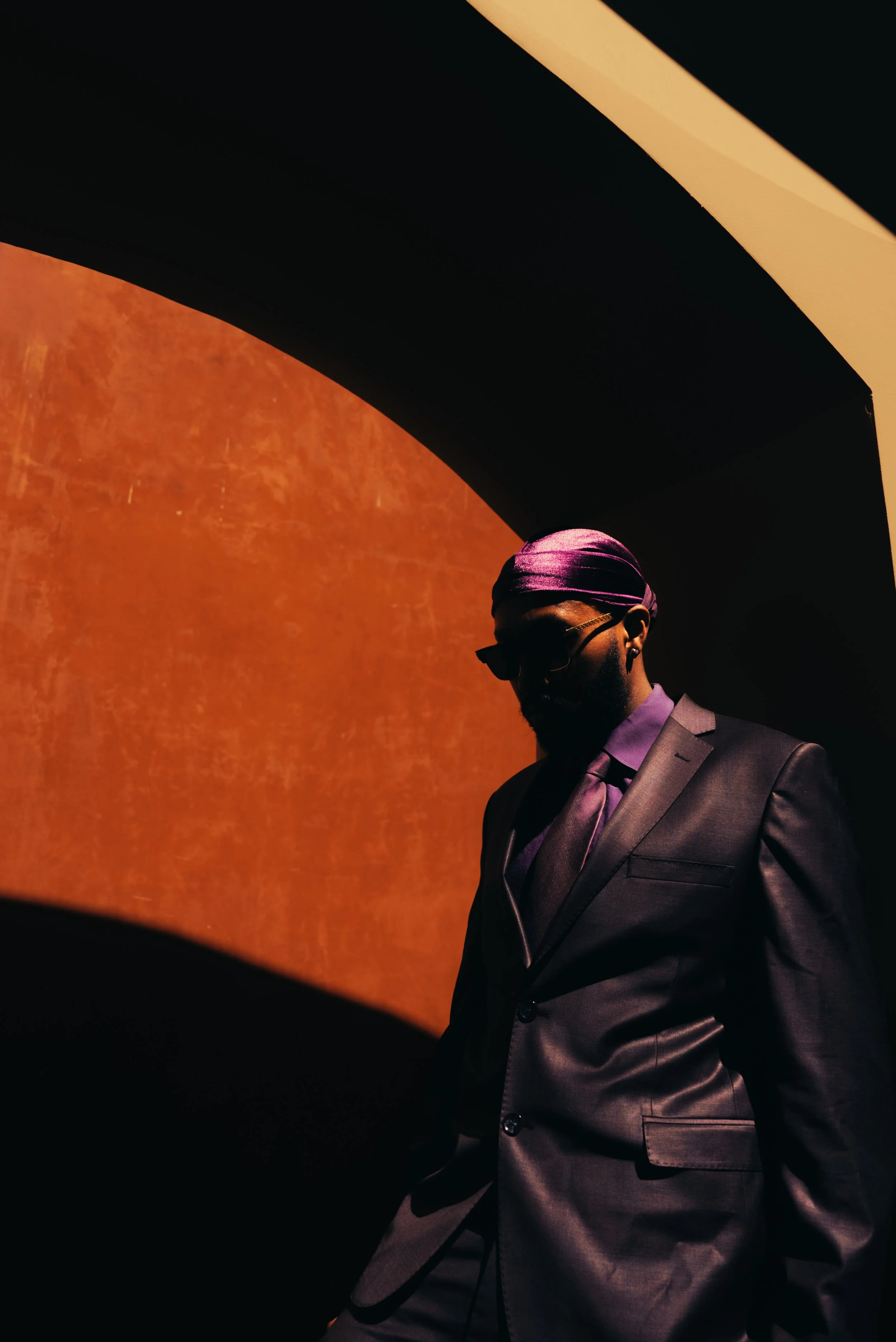 A man in a suit stands in shadows, wearing sunglasses and a purple headscarf, with a large orange-red wall and dark shadows in the background.