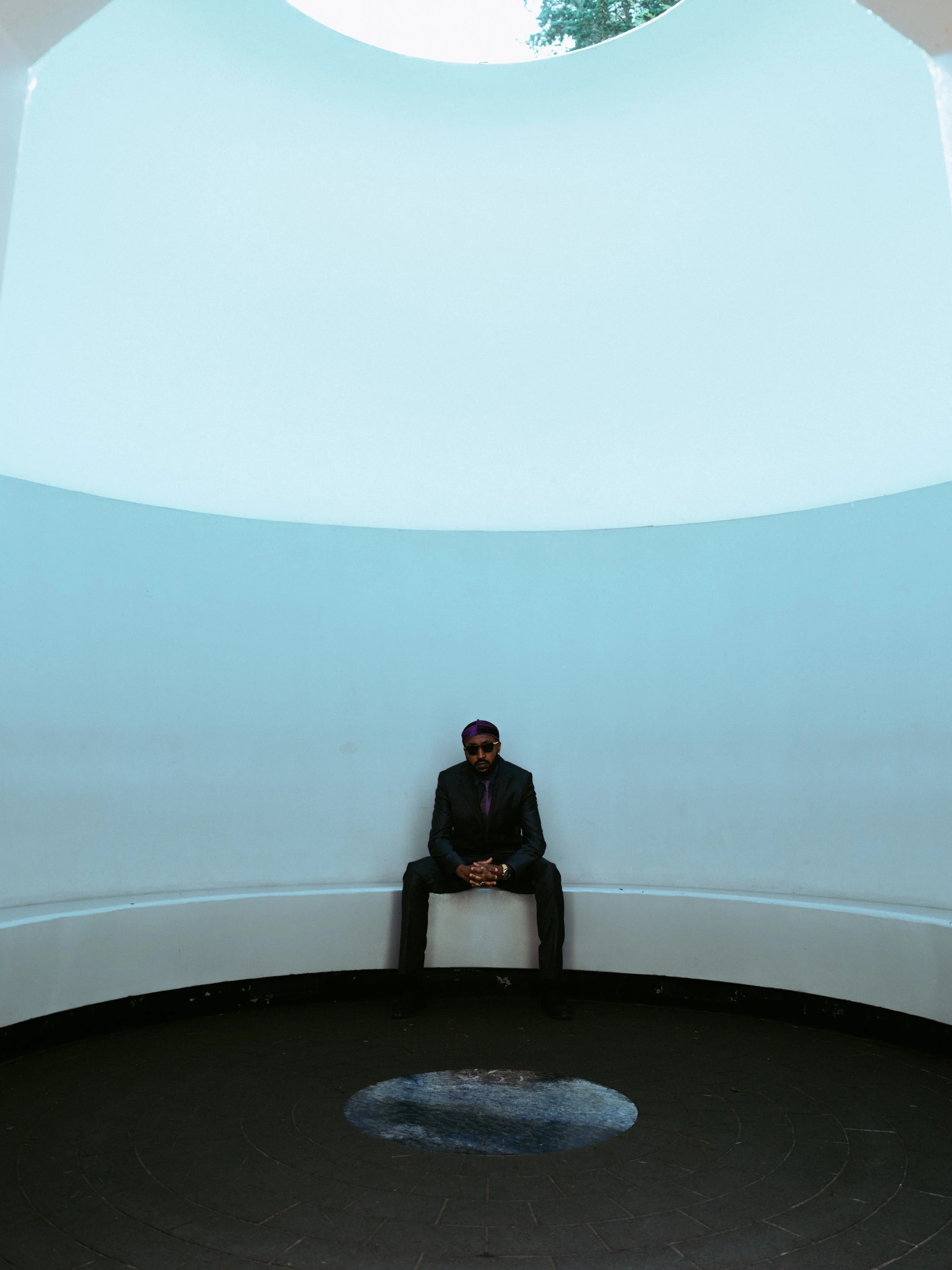 A man wearing sunglasses, a dark suit, and a purple cap sitting on a curved white bench inside a circular structure with a white ceiling and dark flooring.