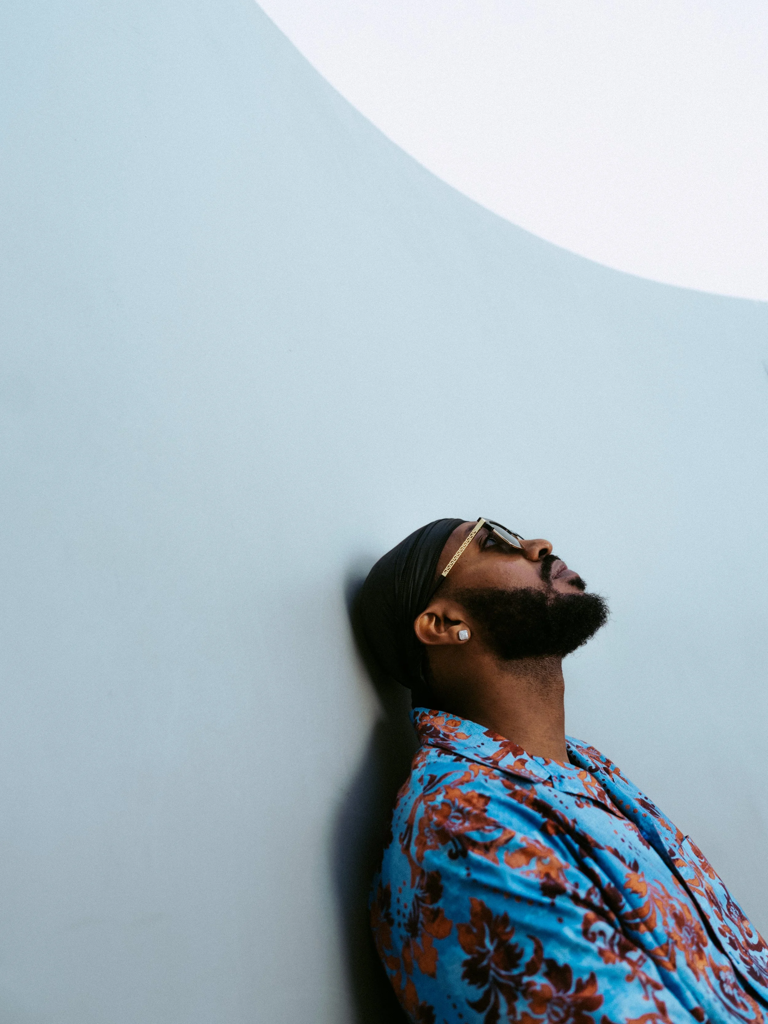A man with a beard, earrings, and sunglasses leaning against a light gray wall, wearing a black headband and a colorful floral shirt.