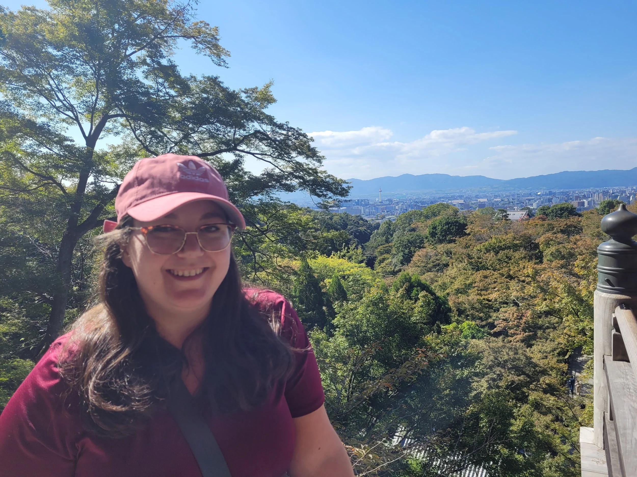 A woman with glasses, long dark hair, a pink cap, and a maroon shirt smiling in a park with green trees, a city skyline, and mountains in the background on a sunny day.