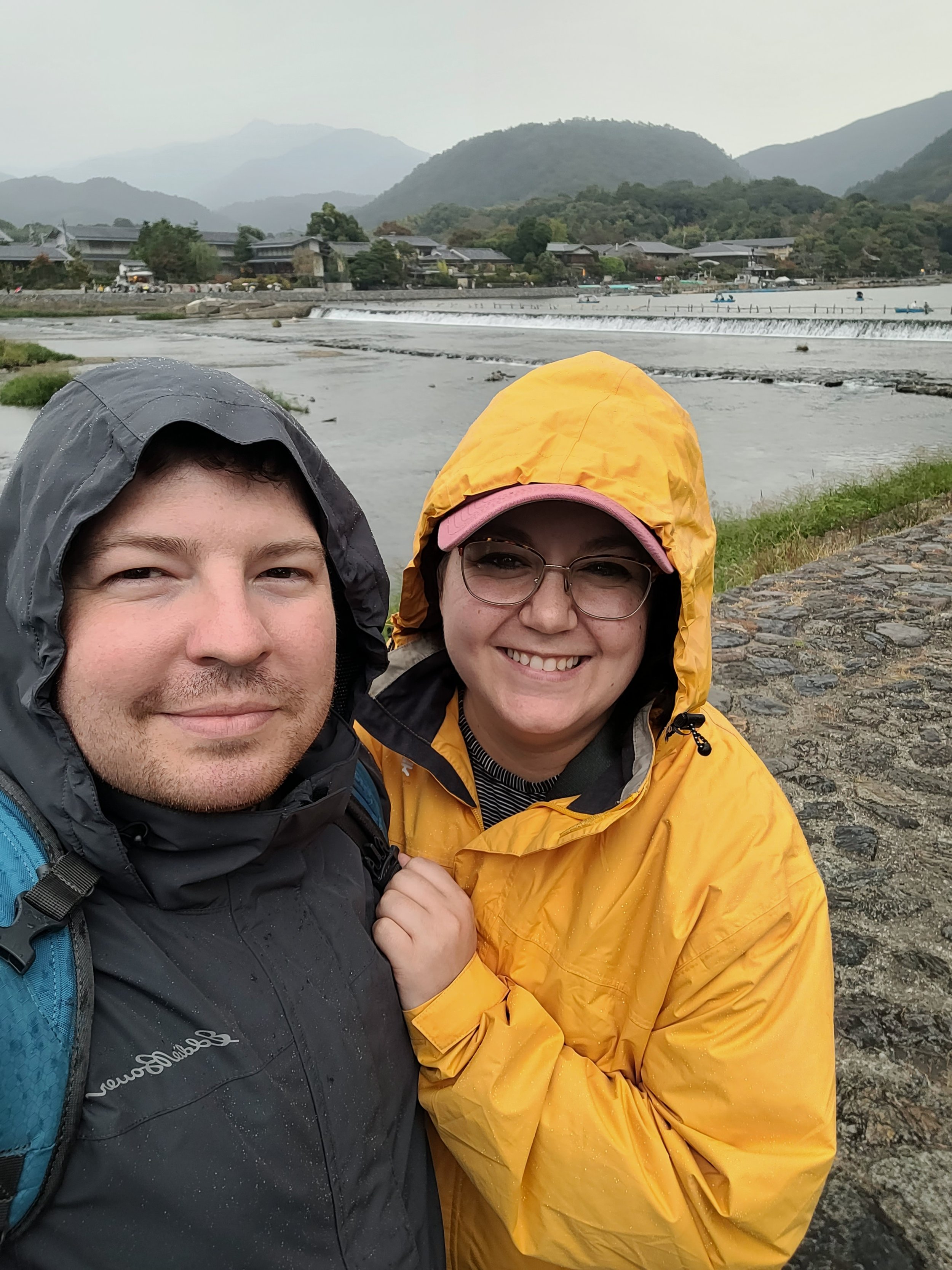A man and woman in rain jackets taking a selfie outdoors with a river, mountains, and traditional buildings in the background on a cloudy day.