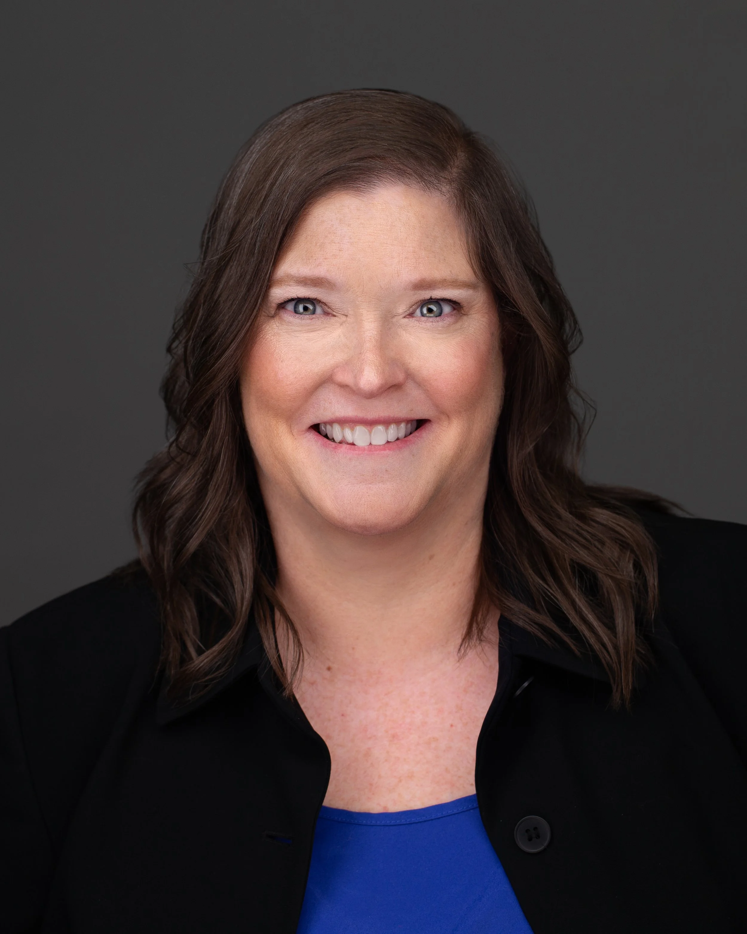 A professional headshot of a smiling woman with shoulder-length brown hair, wearing a black blazer and a blue top, against a dark gray background.