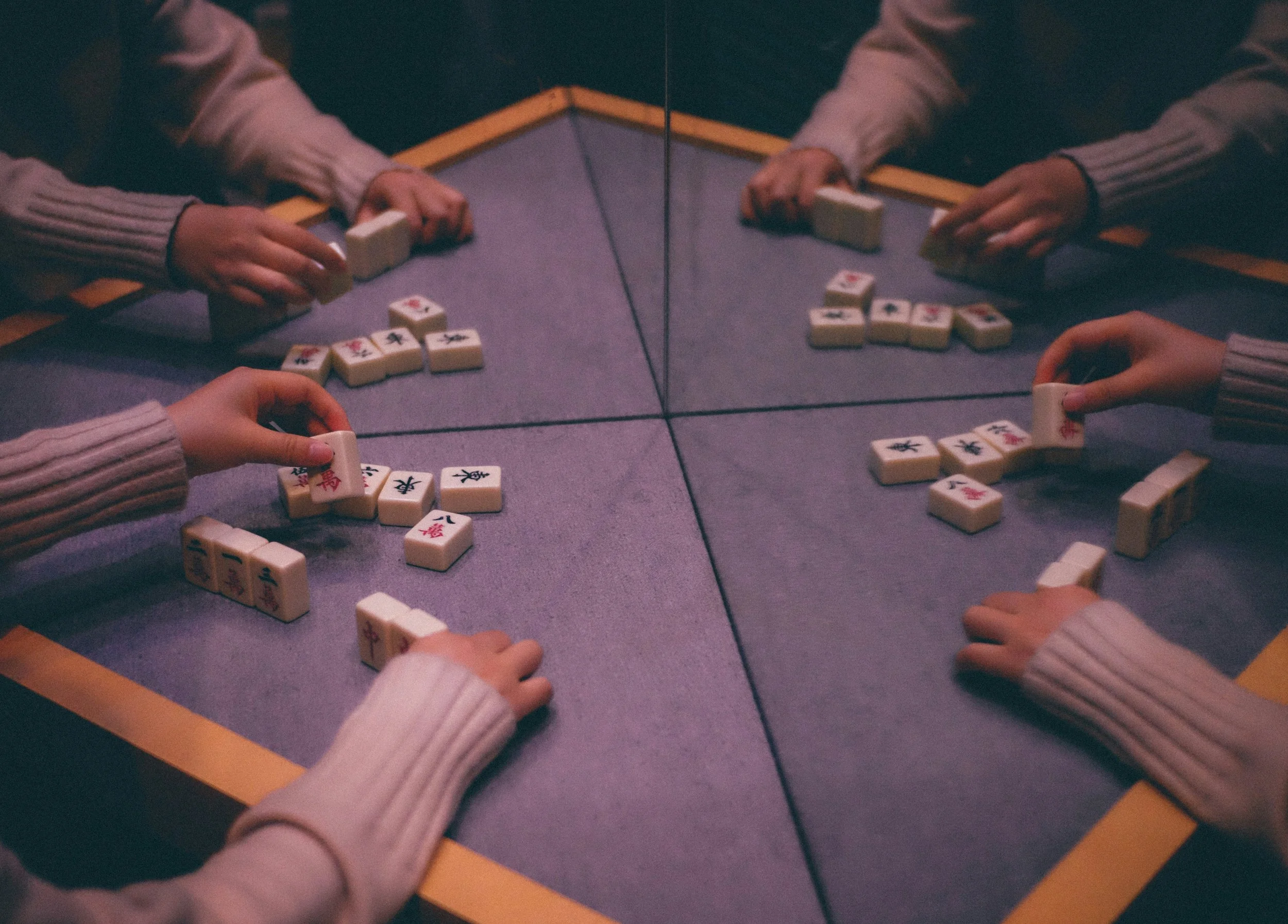 Four people playing Mahjong at a square table with a mirror reflecting their hands and tiles.