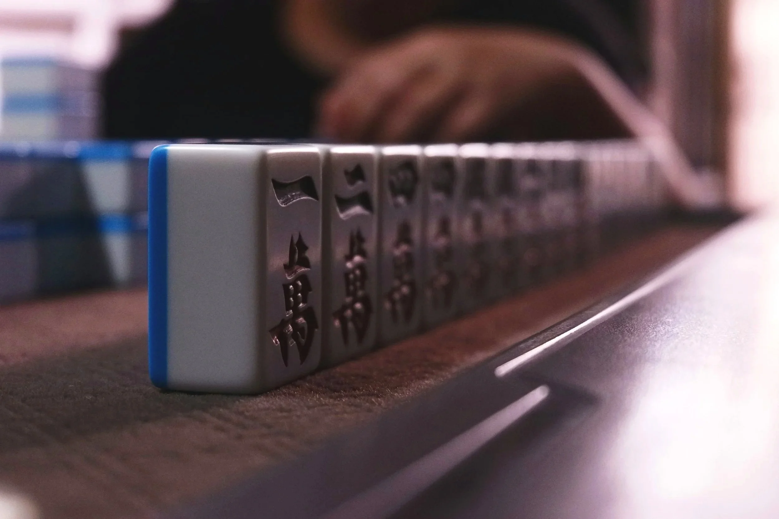Close-up of a mahjong tile with Chinese characters, resting on a wooden surface with a blurred figure in the background.