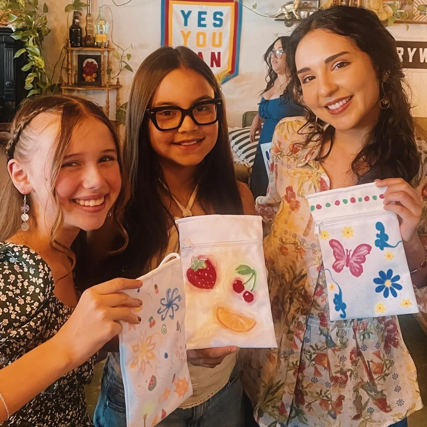 Three women smiling and holding decorated gift bags with colorful designs and drawings of strawberries, butterflies, flowers, and other shapes. They are in a cozy, decorated room with a sign in the background that says 'YES YOU CAN'.