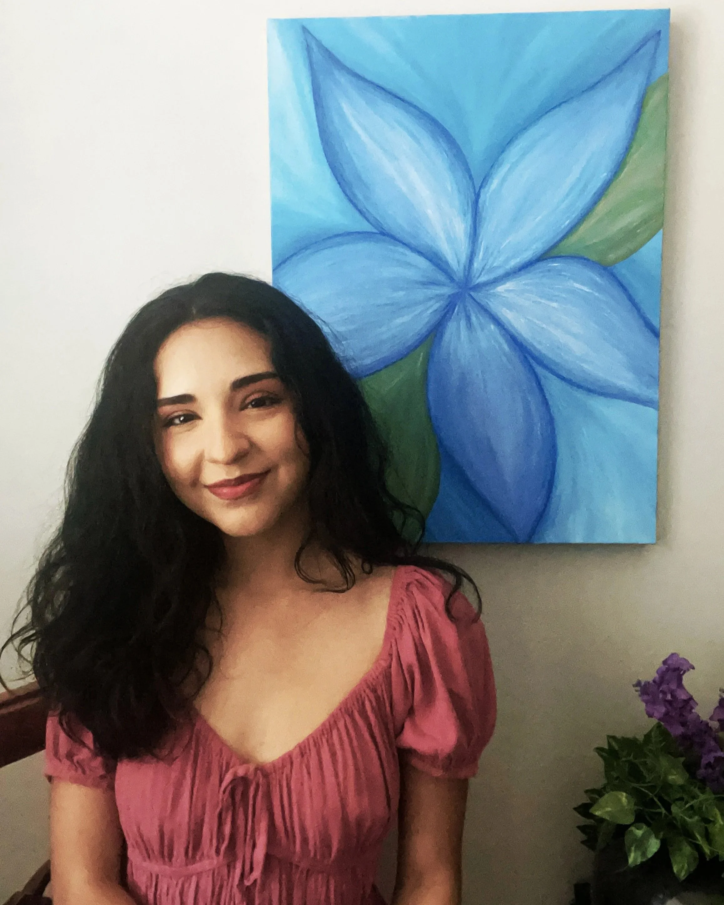 A woman with long dark curly hair smiling in front of a blue flower painting on a white wall, with purple flowers in a pot nearby.