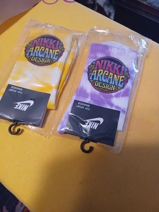 Two Nike socks, one yellow and white and the other purple and white, in plastic packaging on a yellow surface. Each sock features a colorful Nikki Arcane Design logo.