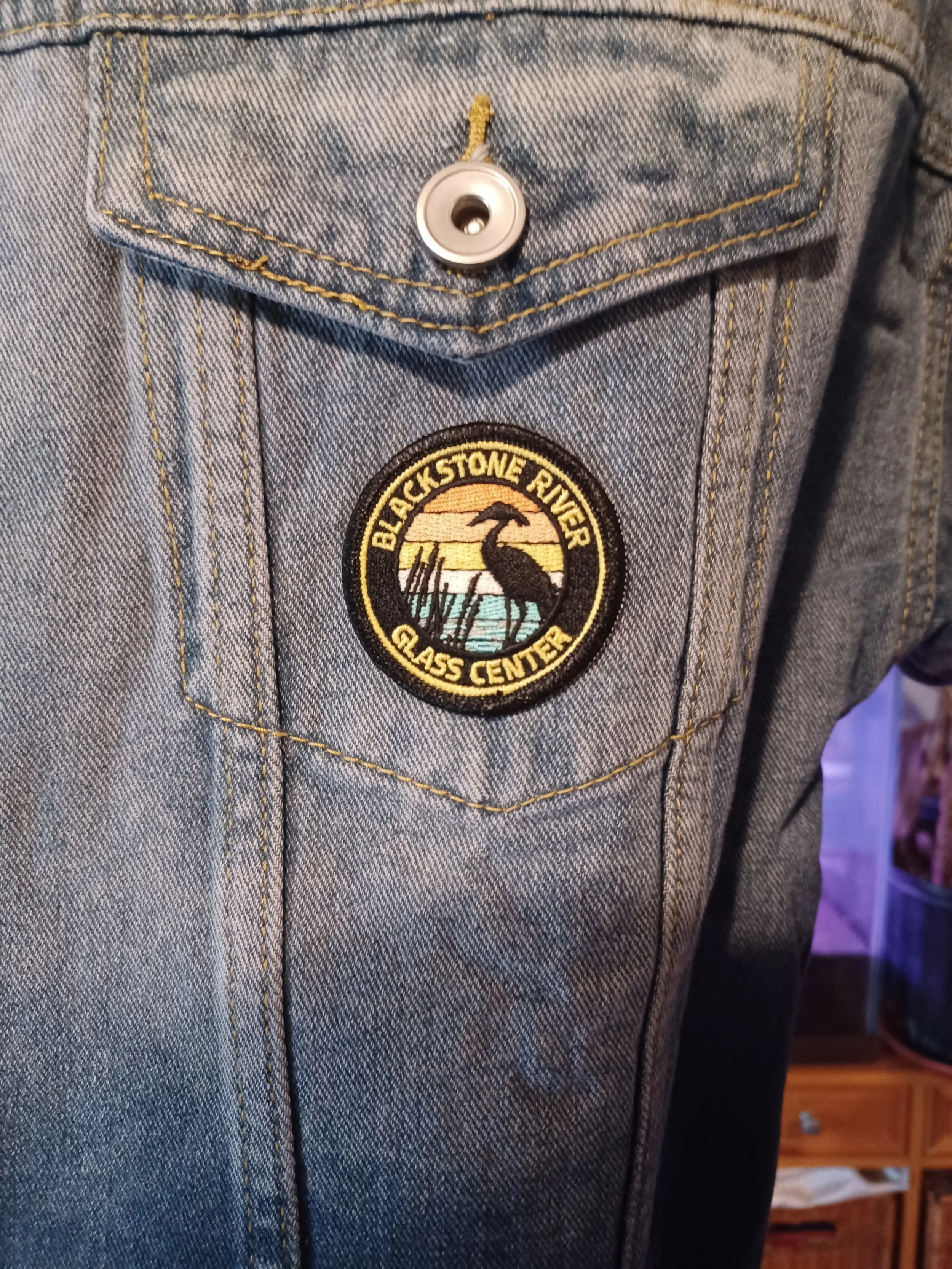 A patch of the Blackstone River Glass Center logo which is a silhouette of a heron and colorful background