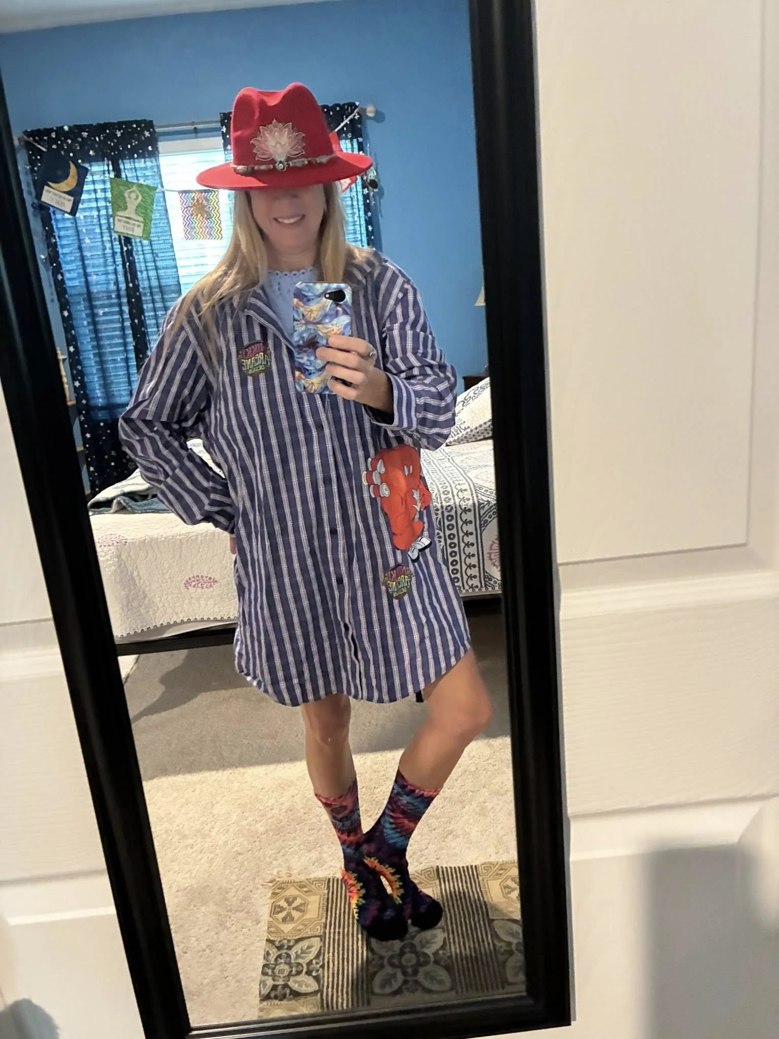 A woman taking a mirror selfie in a bedroom, wearing a striped oversized shirt, a red hat with a large white flower, colorful patterned socks, and holding a phone with a Marvel superhero case.