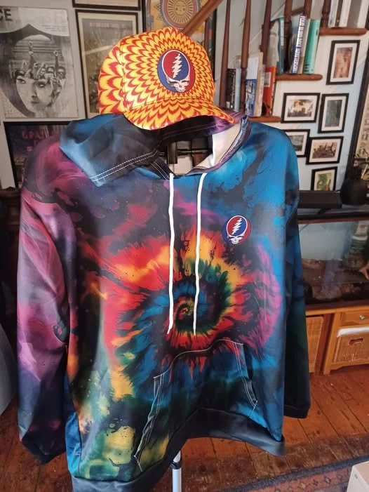 A tie-dye hoodie with a Grateful Dead logo on the chest, and a colorful, patterned baseball cap with the same logo on the front, displayed on a hanger in a room with framed pictures and books in the background.
