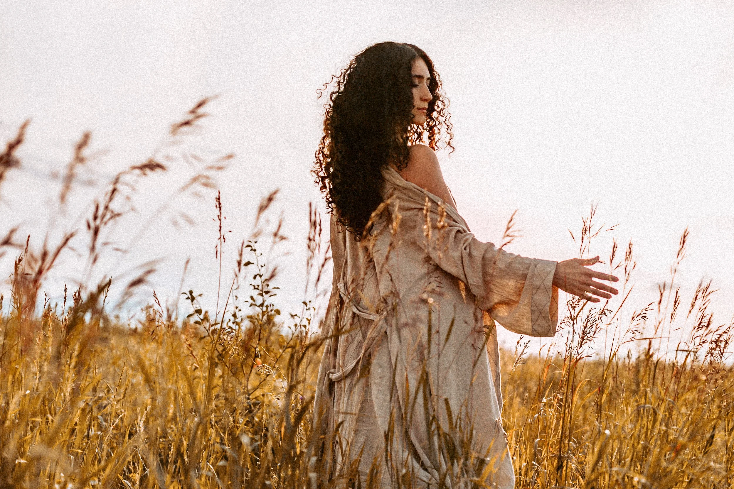 A woman with curly hair standing in a golden wheat field at sunset, gently touching the wheat stalks.