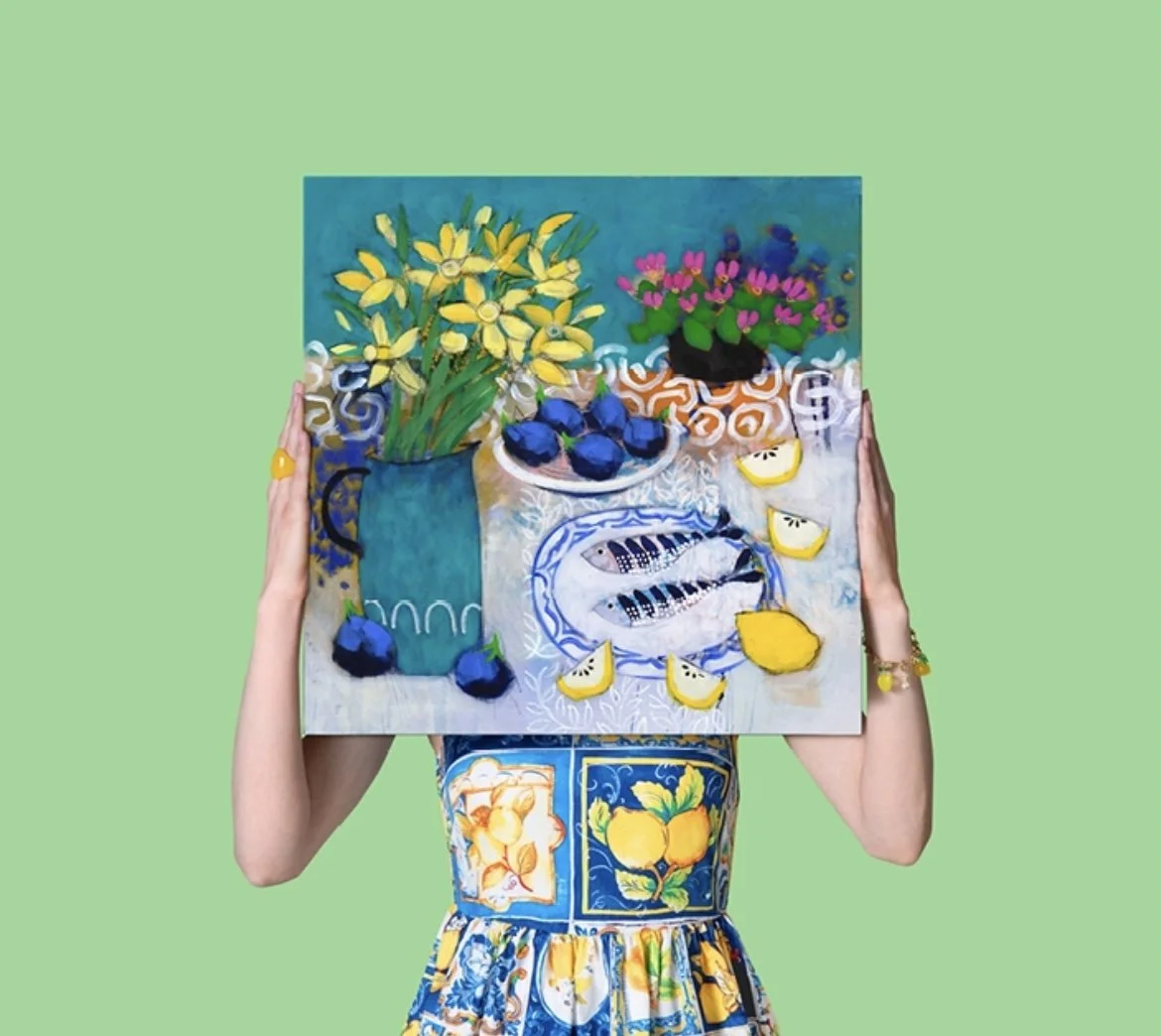Person holding a colorful still life painting featuring yellow and purple flowers in a blue vase, a plate with fish, lemon wedges, and blueberries against a green background.