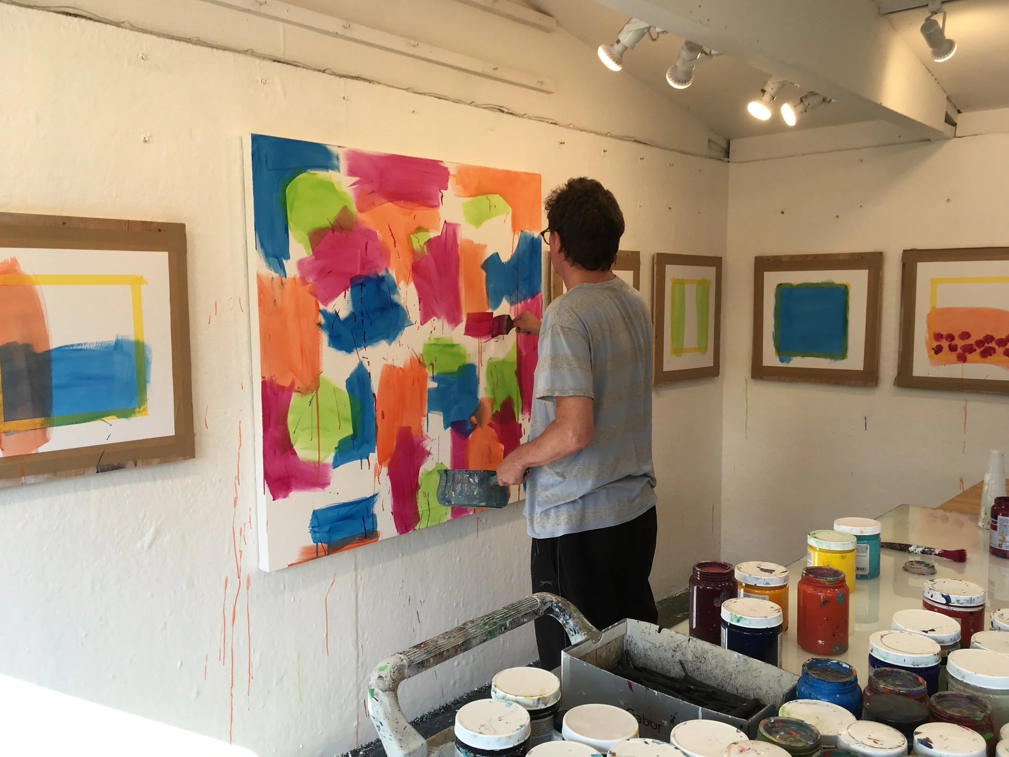 A man with dark curly hair and glasses painting an abstract colorful artwork on a wall in an art gallery, surrounded by several framed paintings and art supplies on a table.