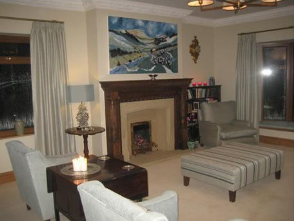 Living room with a fireplace, two windows with curtains, a painting above the fireplace, two chairs, an ottoman, and a side table with a lamp and decorative candles.