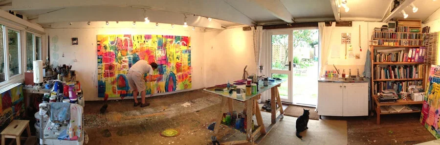 Artist working on a colorful abstract mural inside a well-lit studio with bookshelves, a sliding glass door, and a black cat sitting on the floor.