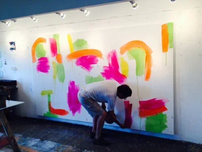 Person painting colorful abstract shapes on a white wall with neon pink, green, orange, and yellow paint.
