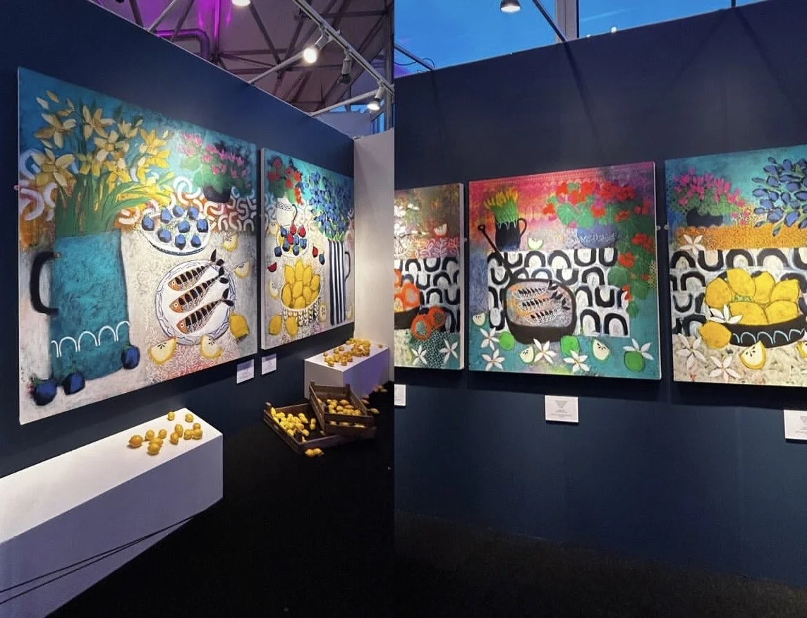 Colorful paintings of vases with flowers, fruits, and fish on a gallery wall, with yellow lemons displayed on white surfaces and in baskets below.